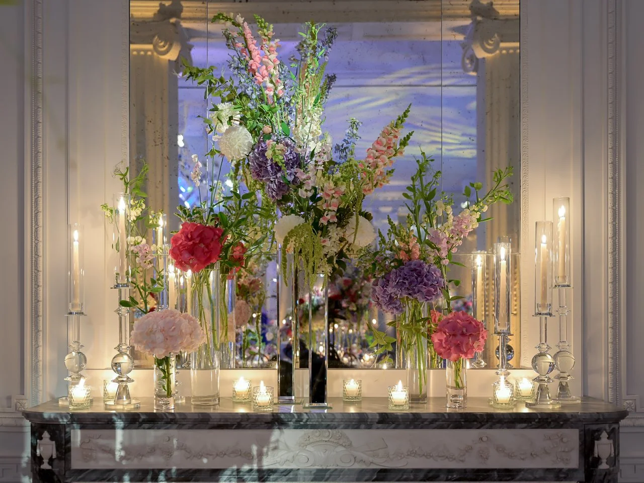 Elegant indoor floral display with pink, purple, and white flowers in glass vases, surrounded by tall candles on a marble table against a mirrored wall.