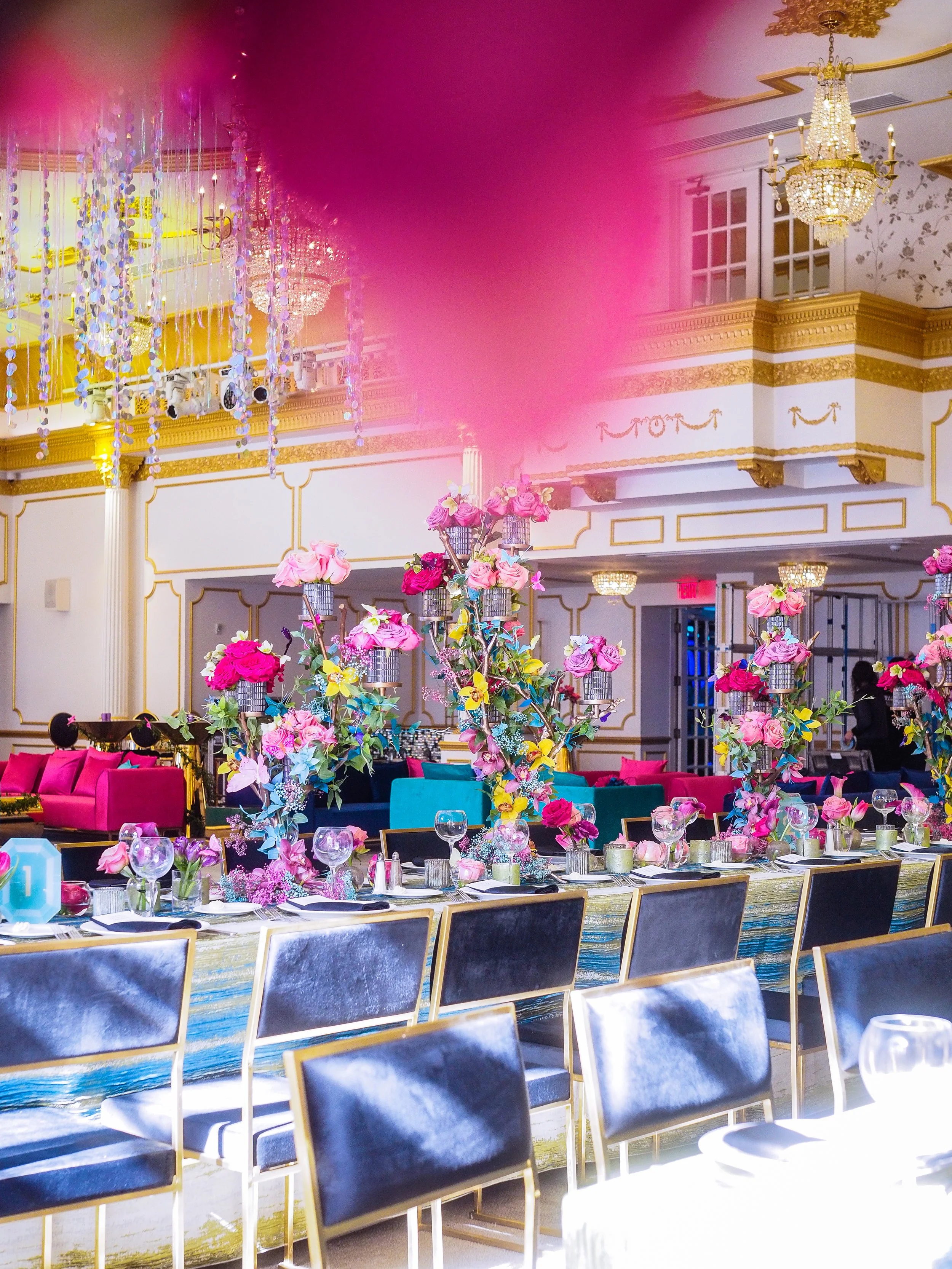Elegant event space decorated with pink and yellow floral centerpieces, crystal chandeliers, and ornate gold-trimmed decor.
