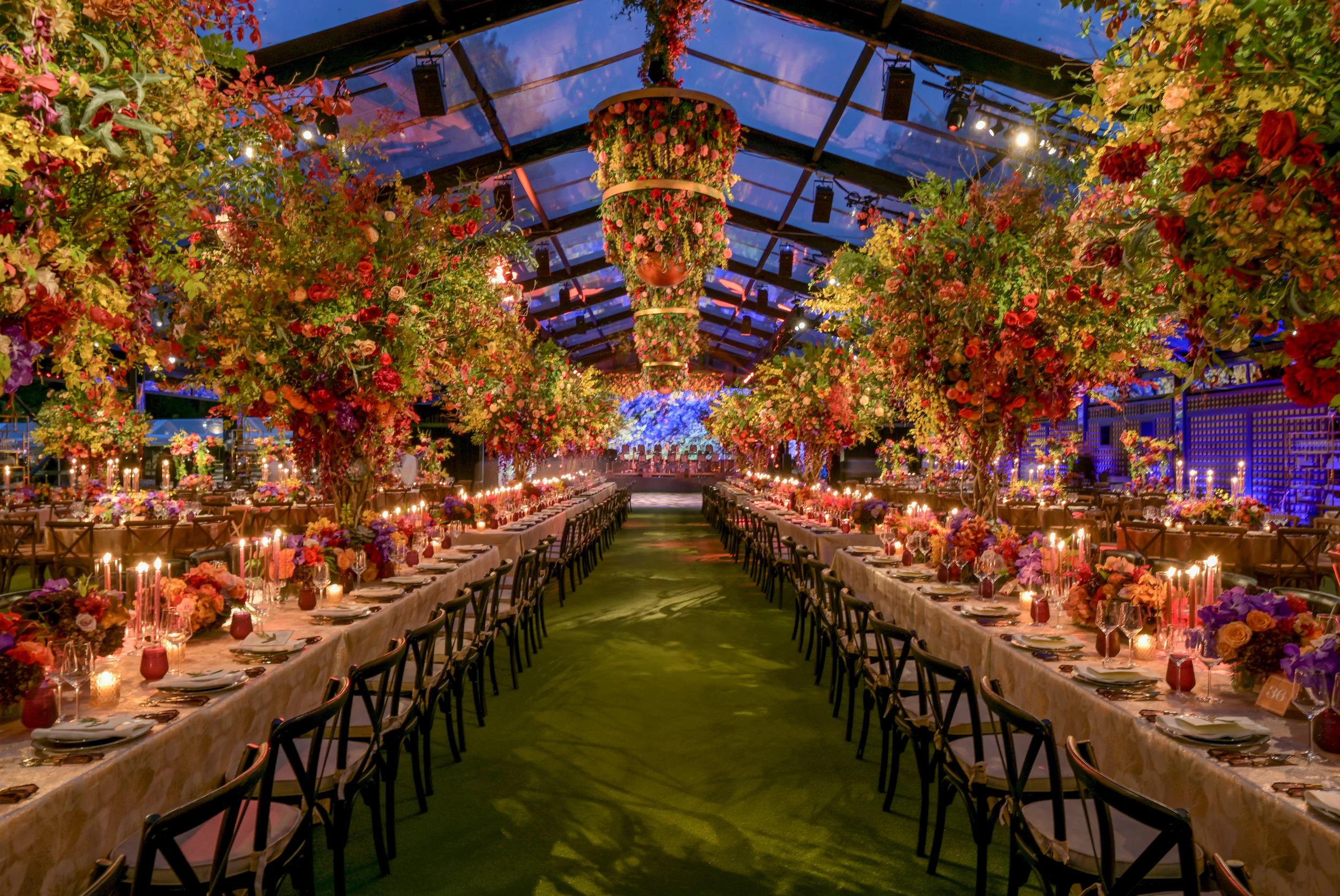A decorated event space with long dining tables set with plates, glasses, and cutlery, surrounded by floral centerpieces and tall floral trees, under a glass ceiling with hanging lights and floral arrangements.