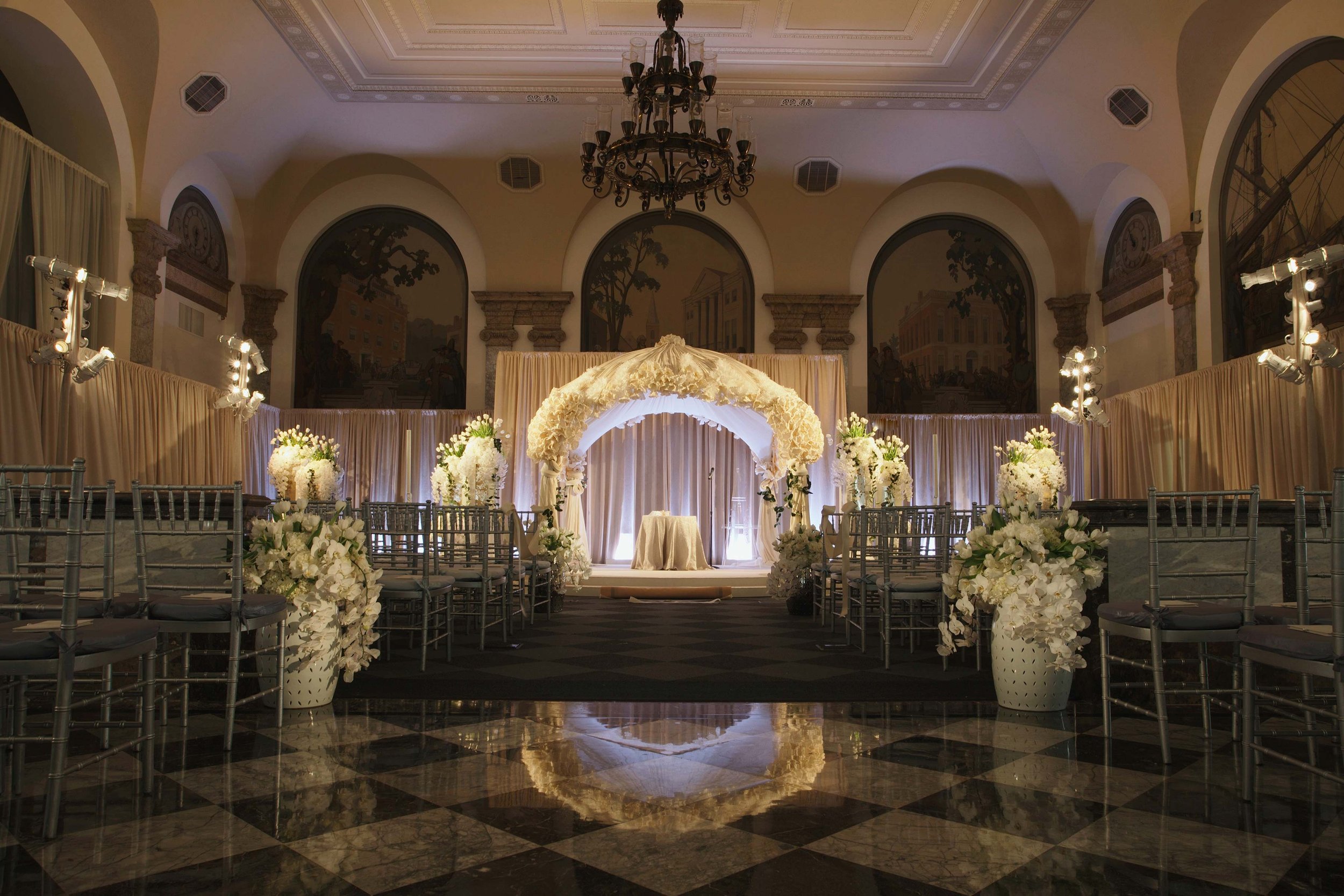 Elegant indoor wedding ceremony setup with white flowers and draped fabric under a floral arch on a stage, with chairs arranged on both sides and chandeliers hanging from the ceiling.