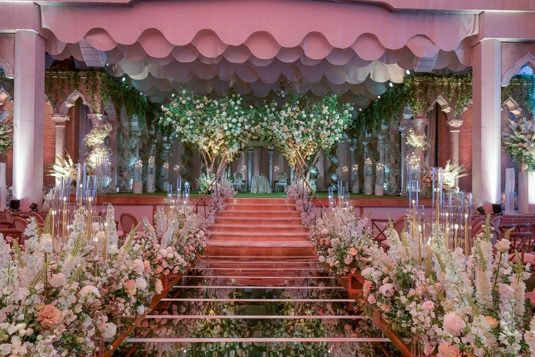 Elegant indoor wedding ceremony setup with pink and white floral arrangements, decorated stairs, and draped ceiling.