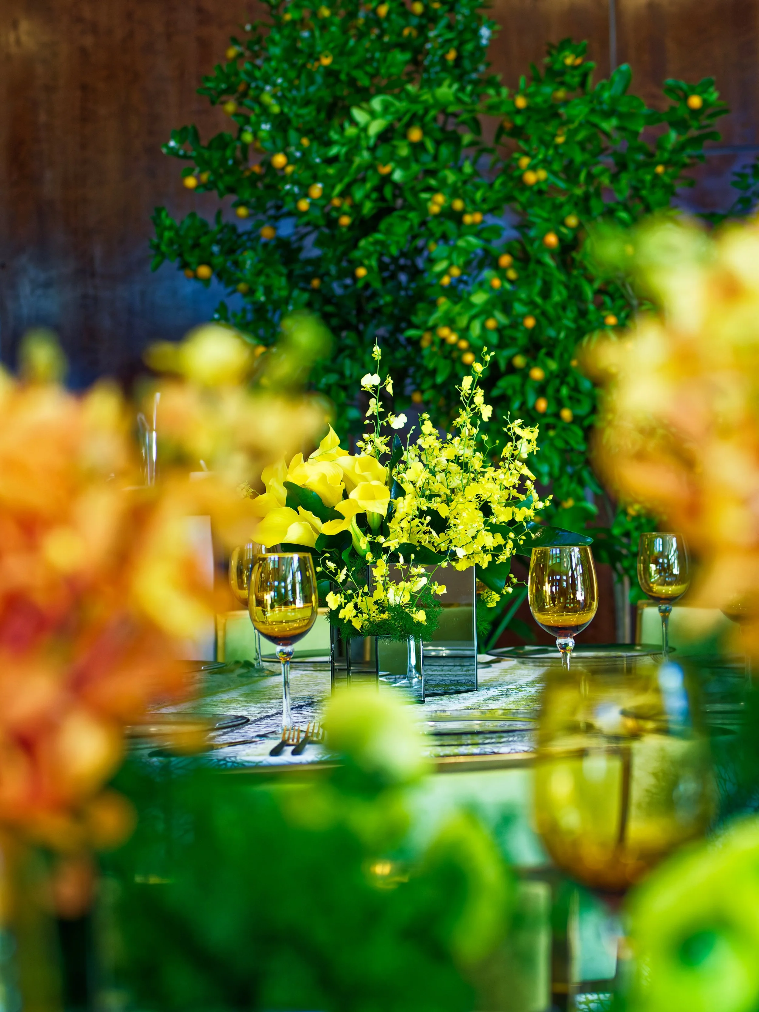 A dining table decorated with yellow flowers and wine glasses, set against a backdrop of a large green citrus tree with yellow fruit and a wooden wall.