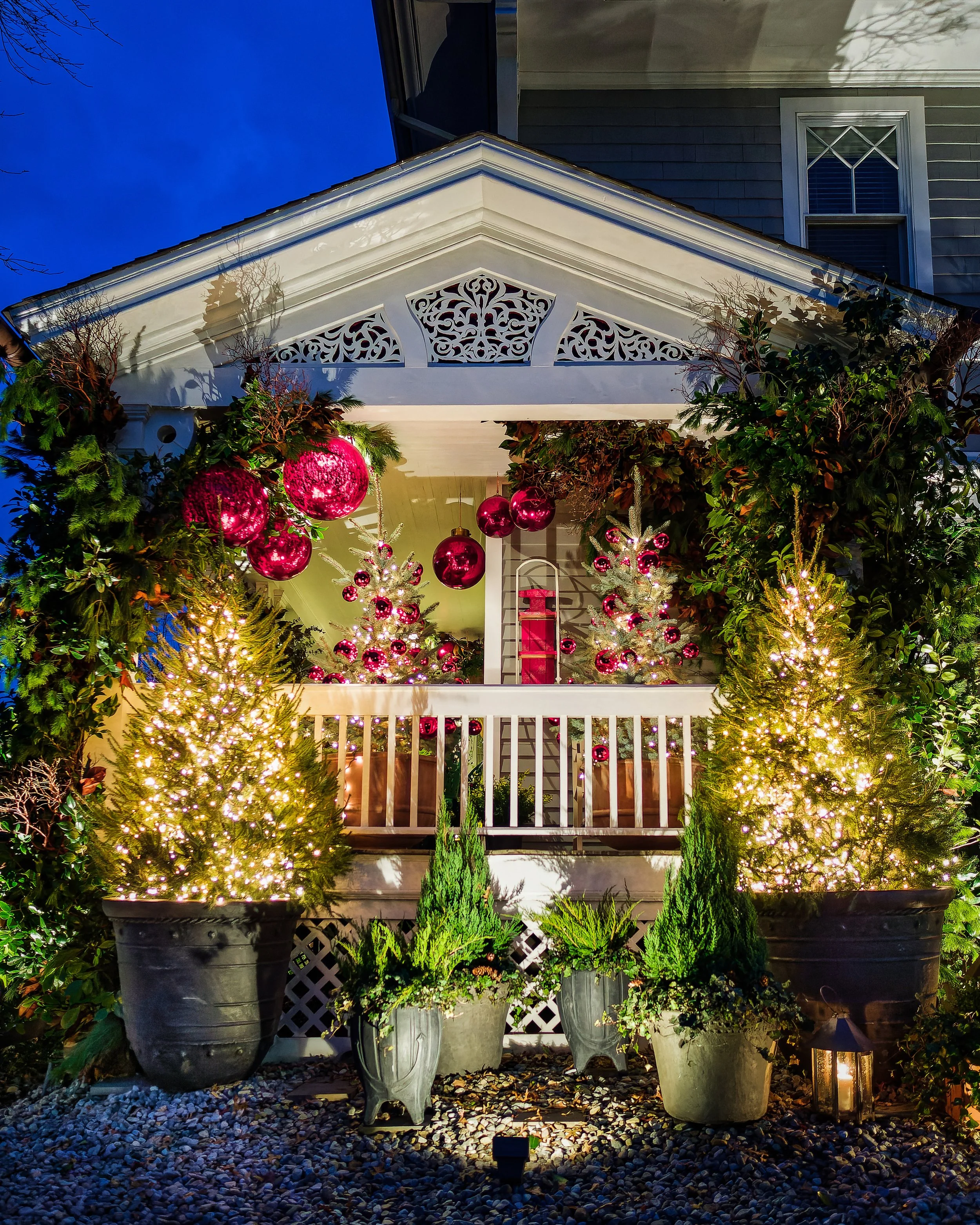 Decorated front porch with illuminated Christmas trees, pink ornaments hanging from a string, and various potted plants, with party decorations inside visible from the porch.