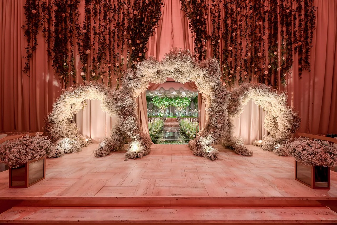 A pink-themed wedding altar decorated with large white flower arrangements, greenery, and draped pink curtains, leading to an outdoor garden setting.