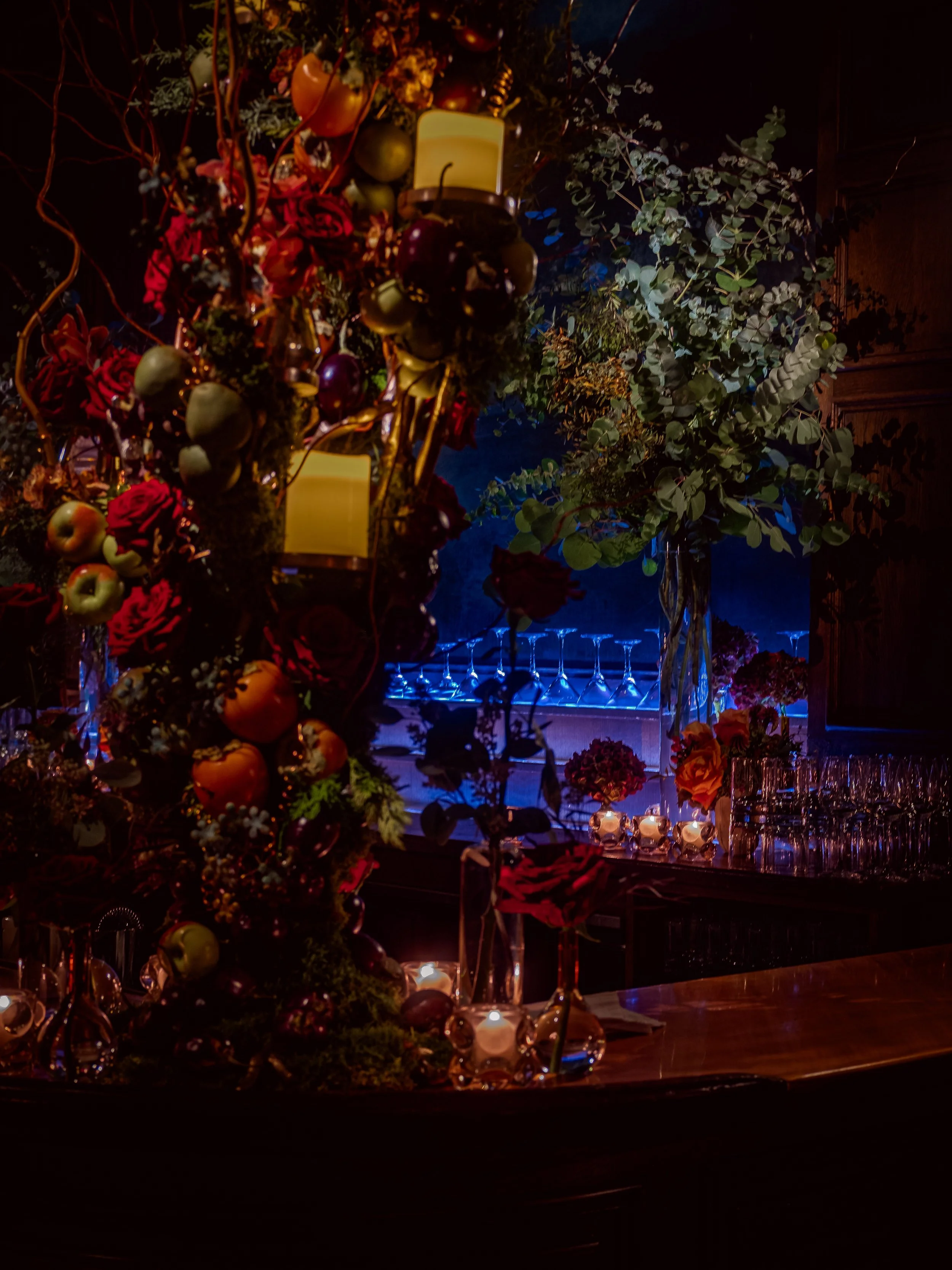 Elegant bar or dining area with colorful floral and fruit arrangements, candles, and glassware, illuminated by warm lighting.