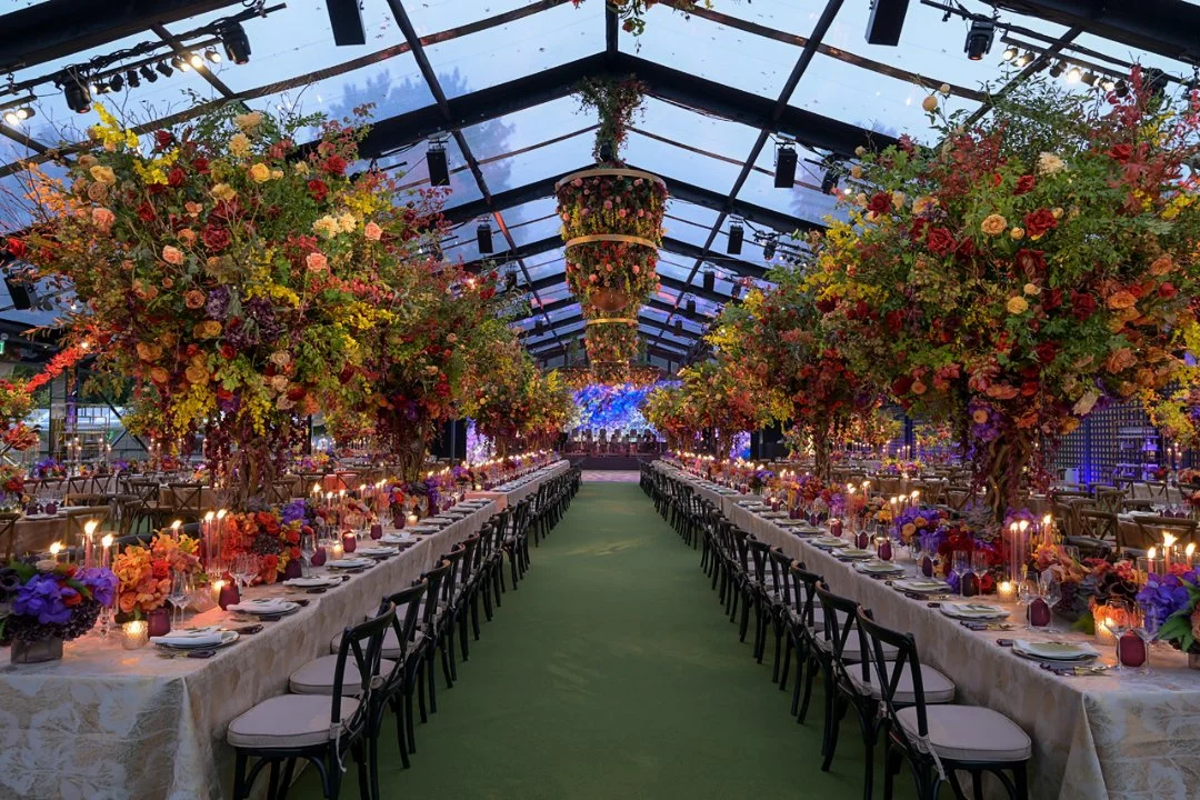 Elegant indoor event space with long banquet tables adorned with colorful floral arrangements and candles, under a glass ceiling with hanging lights.