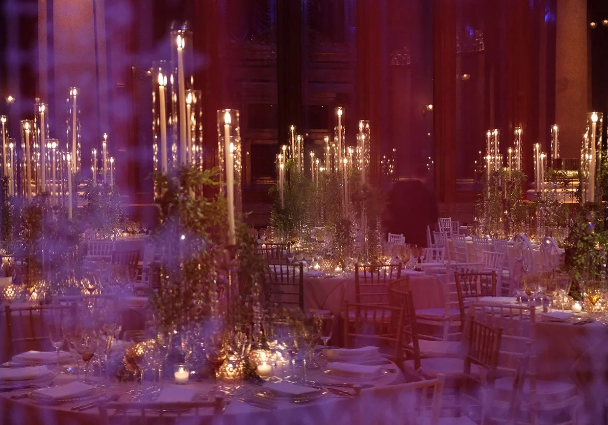 Elegant banquet hall decorated with tall candles, floral centerpieces, and set dining tables with white and gold accents, suitable for a formal event or wedding reception.