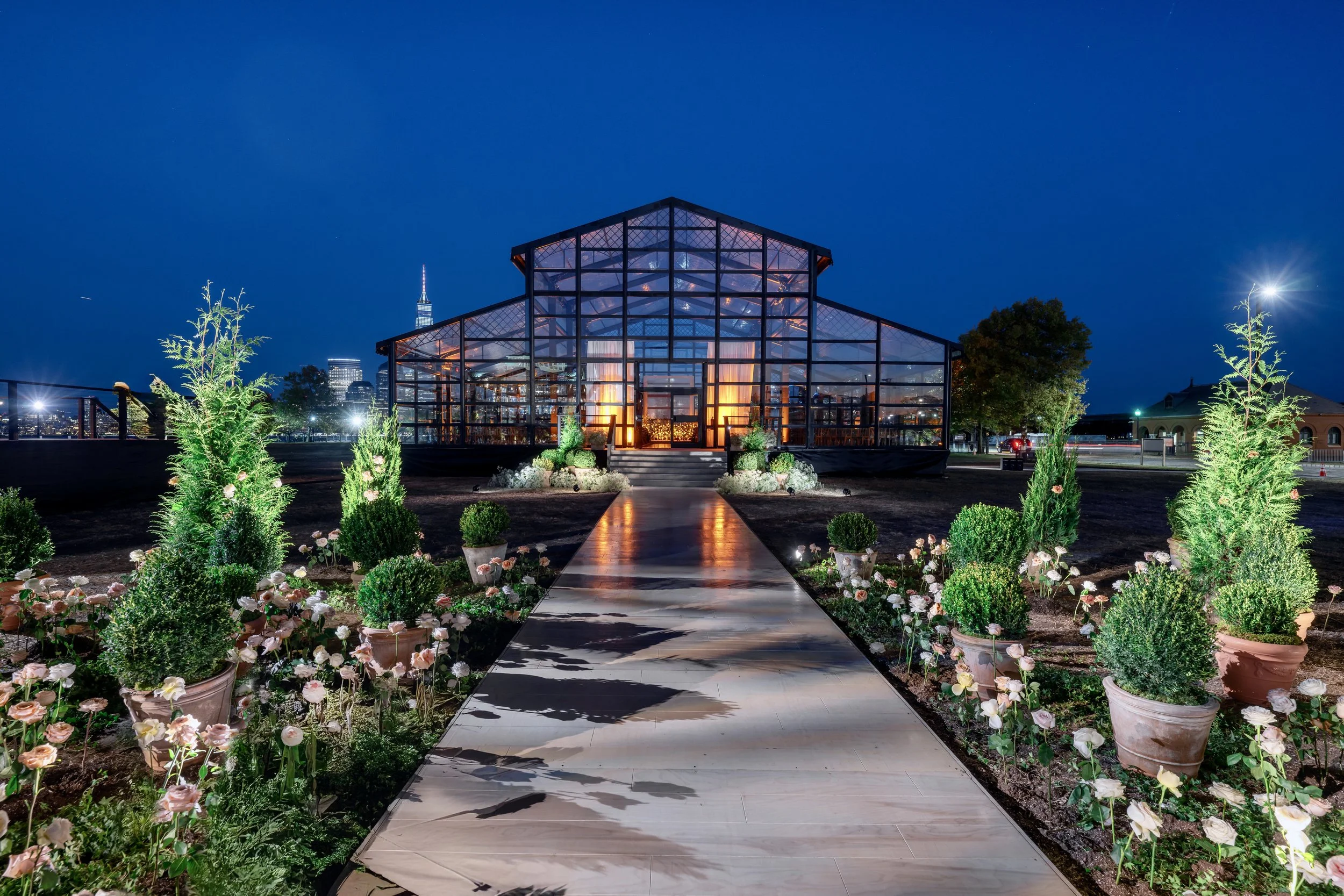 Night view of a glass greenhouse with a lit interior, a pathway leading to the entrance, and potted plants and flowers on either side of the path.