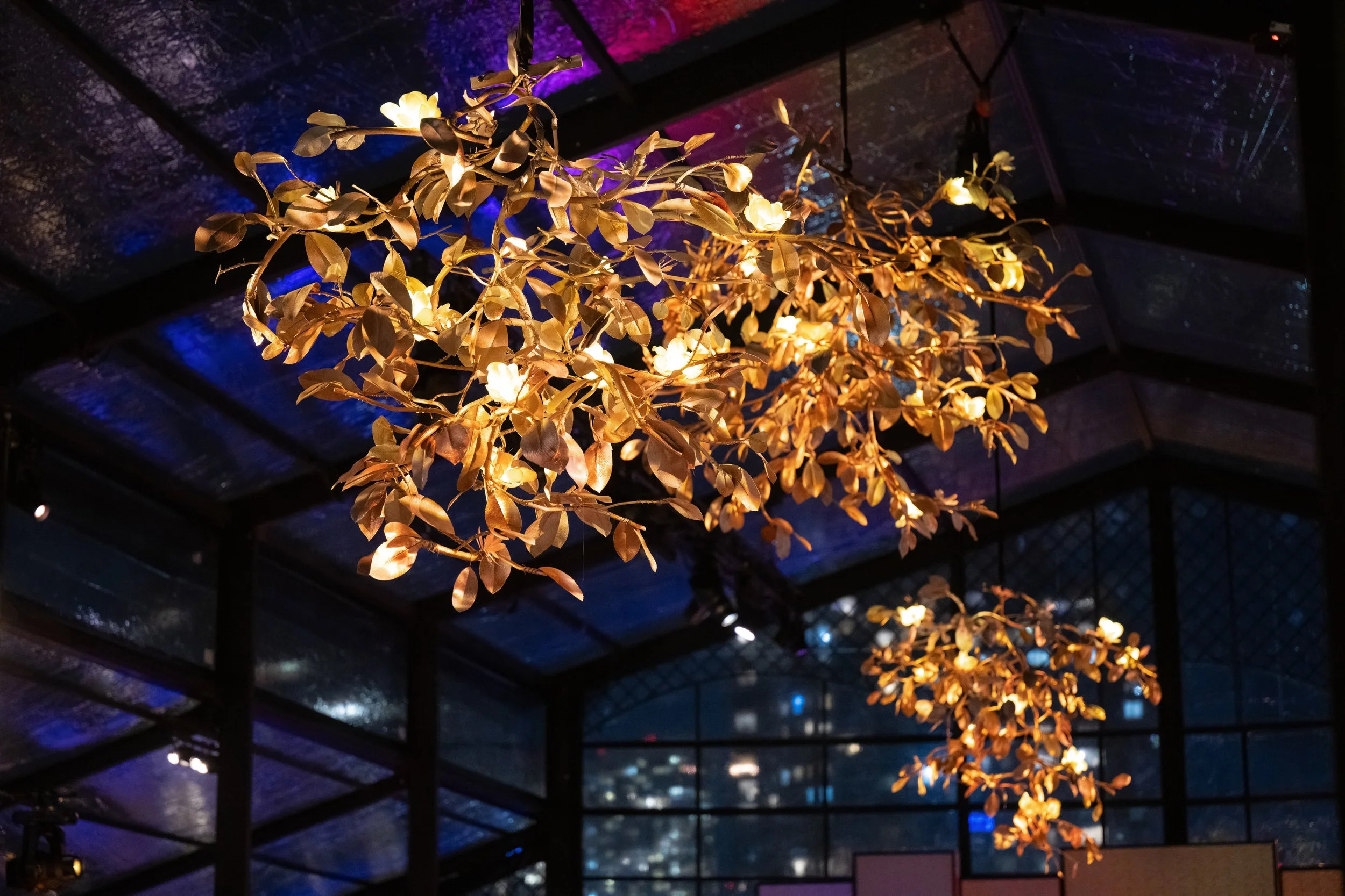Golden leaf and flower chandelier hanging from ceiling, illuminated against a dark backdrop with colorful accent lights.