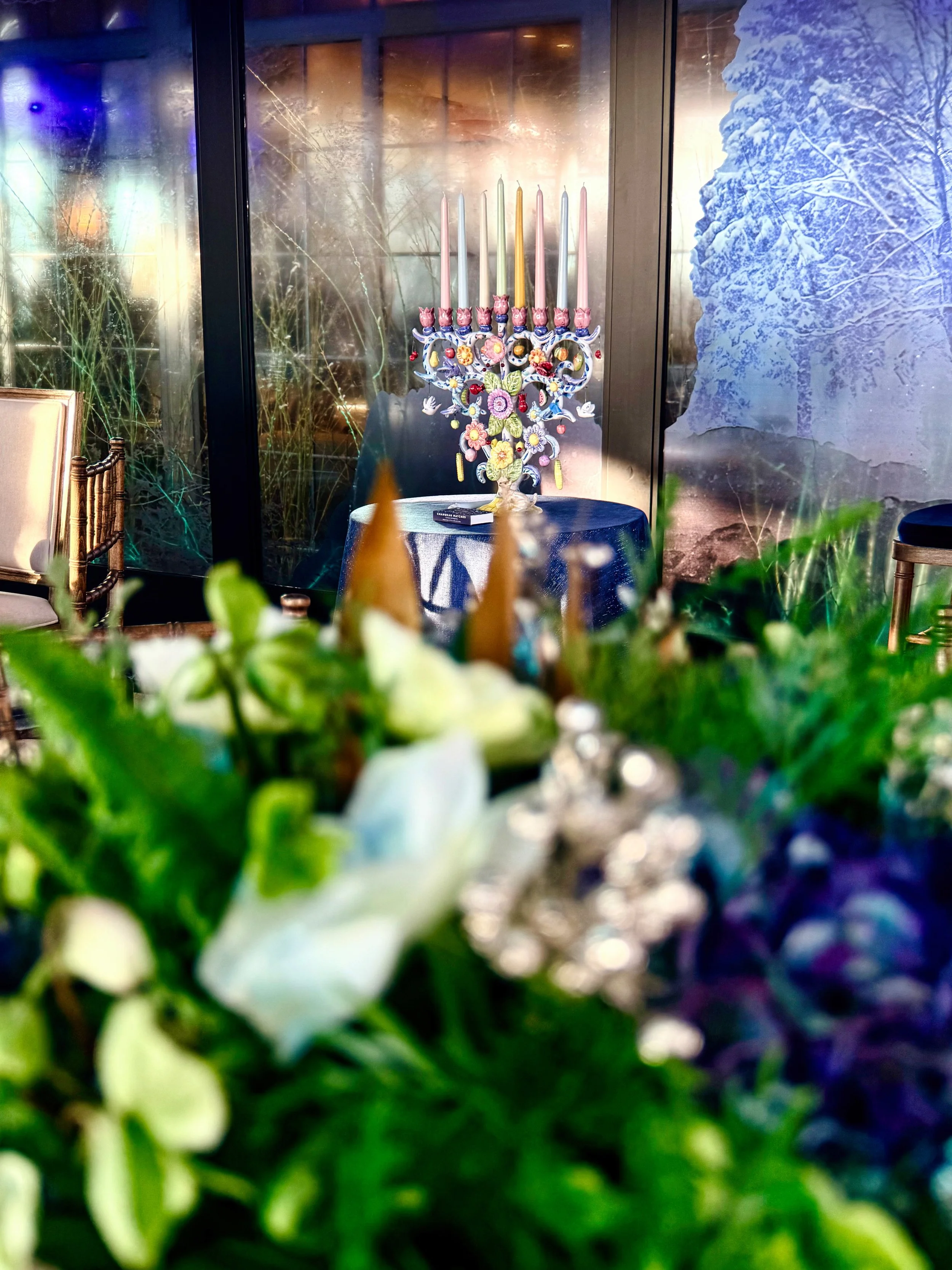 A decorative menorah with colorful candles and floral embellishments on a table, with a wintery landscape and snow-covered trees visible through the windows in the background.