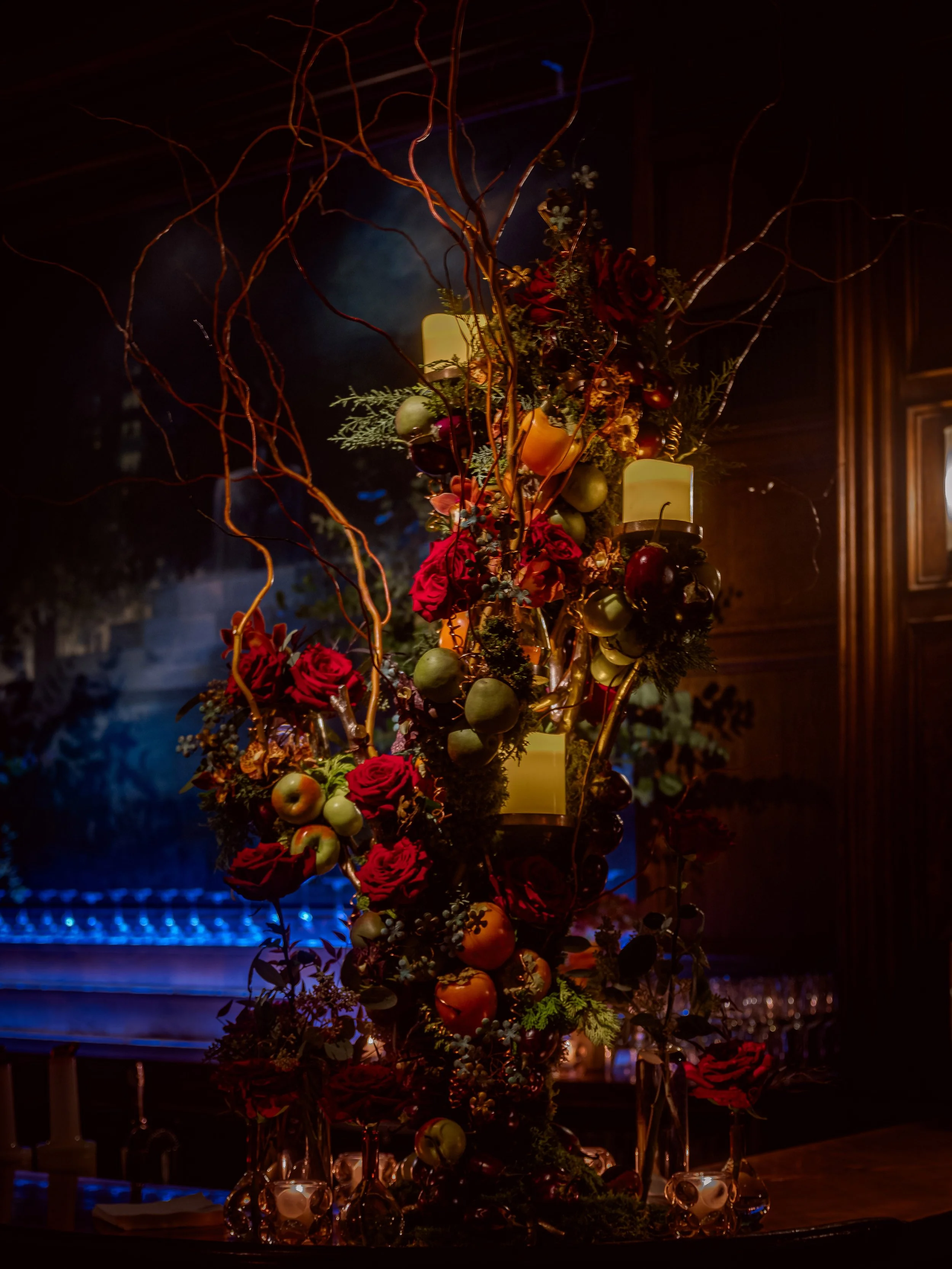 A floral centerpiece with red roses, green apples, and candles arranged on a table in a dimly lit room.