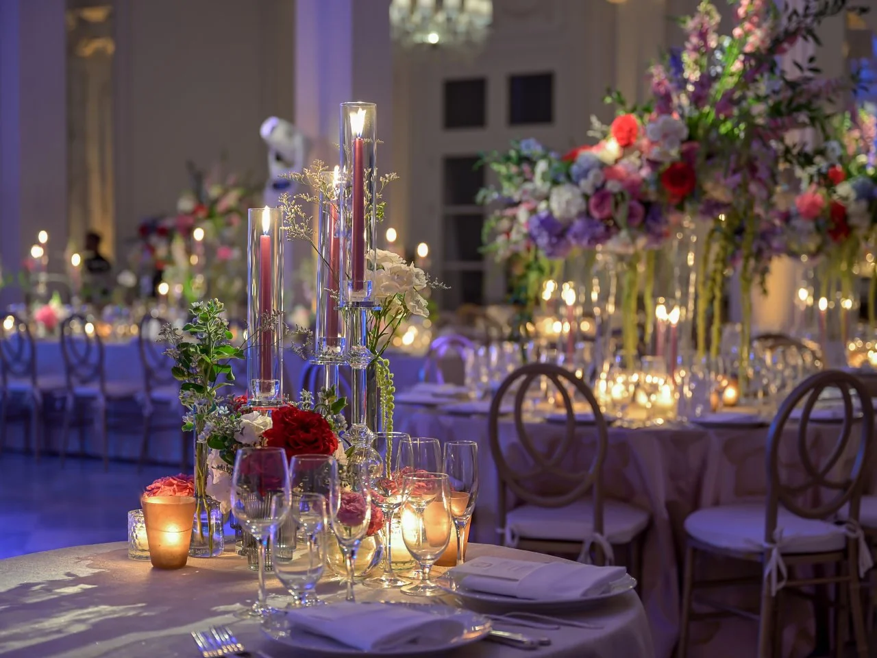 Elegant banquet table decorated with tall glass candle holders, floral centerpieces with roses and other flowers, glassware, plates, and napkins, in a softly lit ballroom with additional tables and floral arrangements in the background.