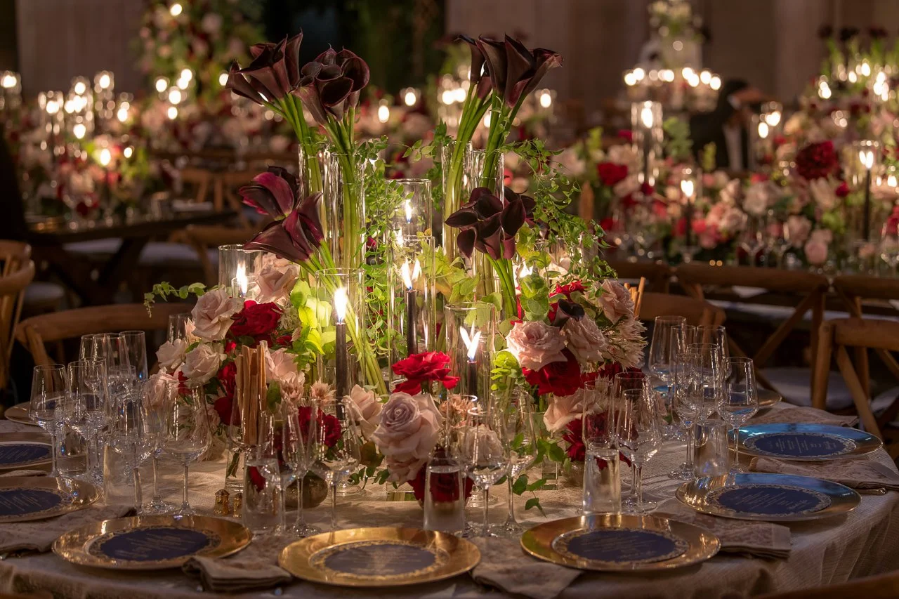 Elegant banquet table decorated with tall dark calla lilies and white and red roses, surrounded by multiple glasses, gold-rimmed plates, and lit candles, in a warmly lit decorated venue.