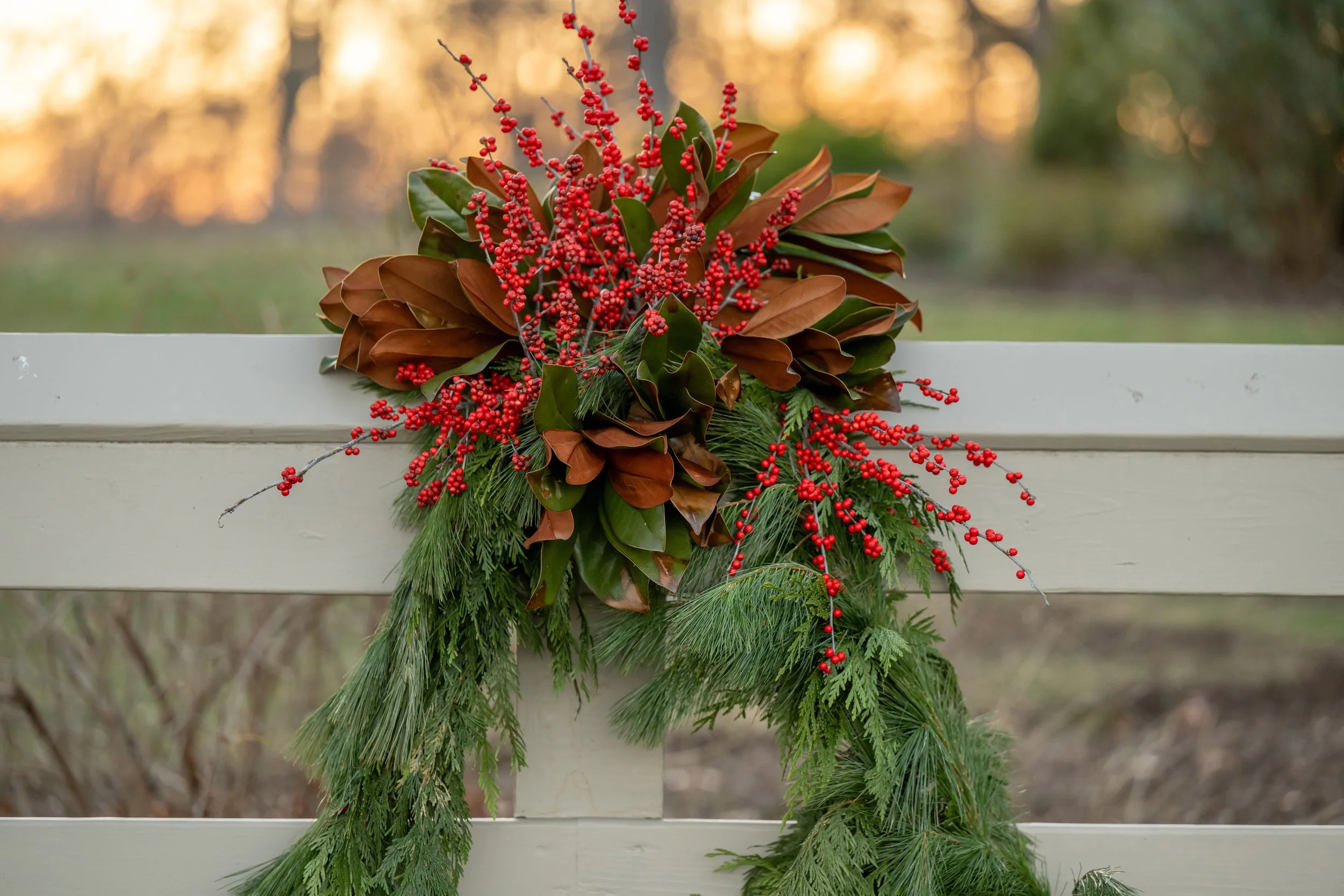 A festive Christmas wreath with red berries, green pine branches, and brown magnolia leaves hanging on a white wooden fence