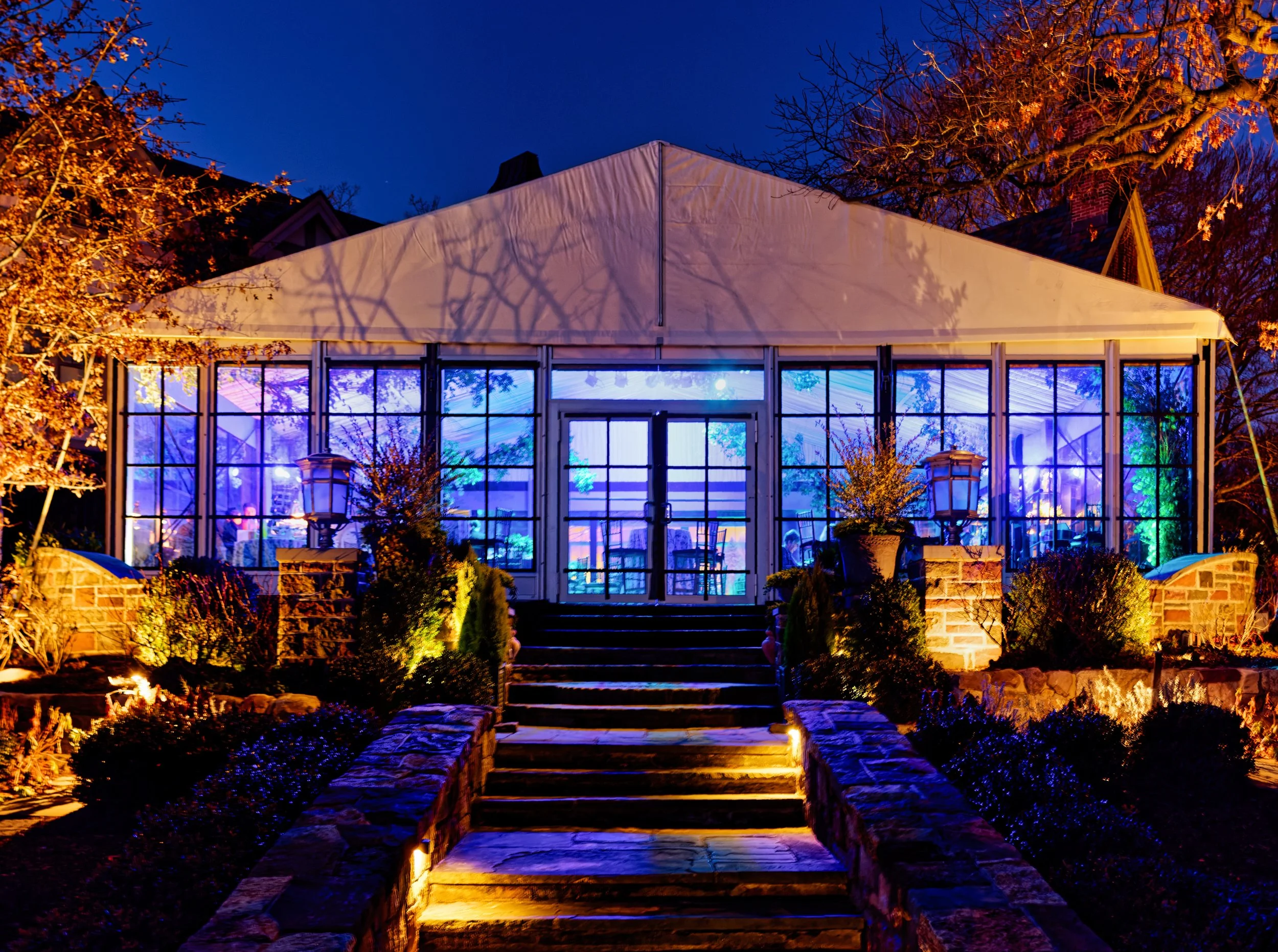 Night view of a glass greenhouse with interior and exterior lighting, stone steps leading to the entrance, surrounded by landscaped bushes and trees.