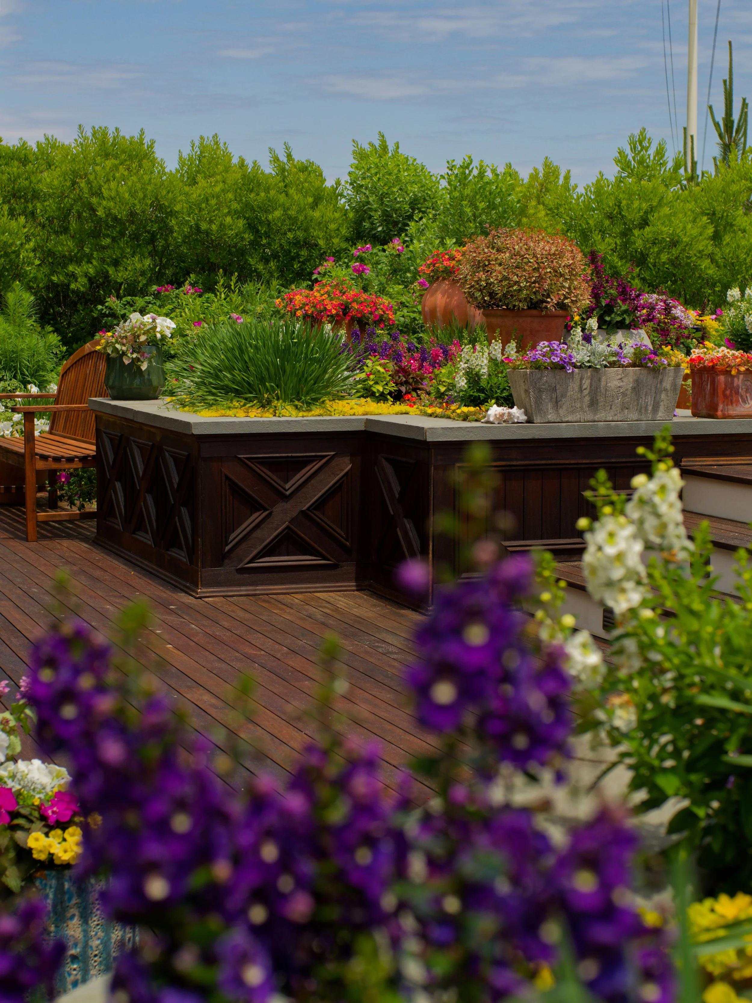 A wooden deck with various colorful potted flowers and plants, surrounded by lush greenery and a clear sky.