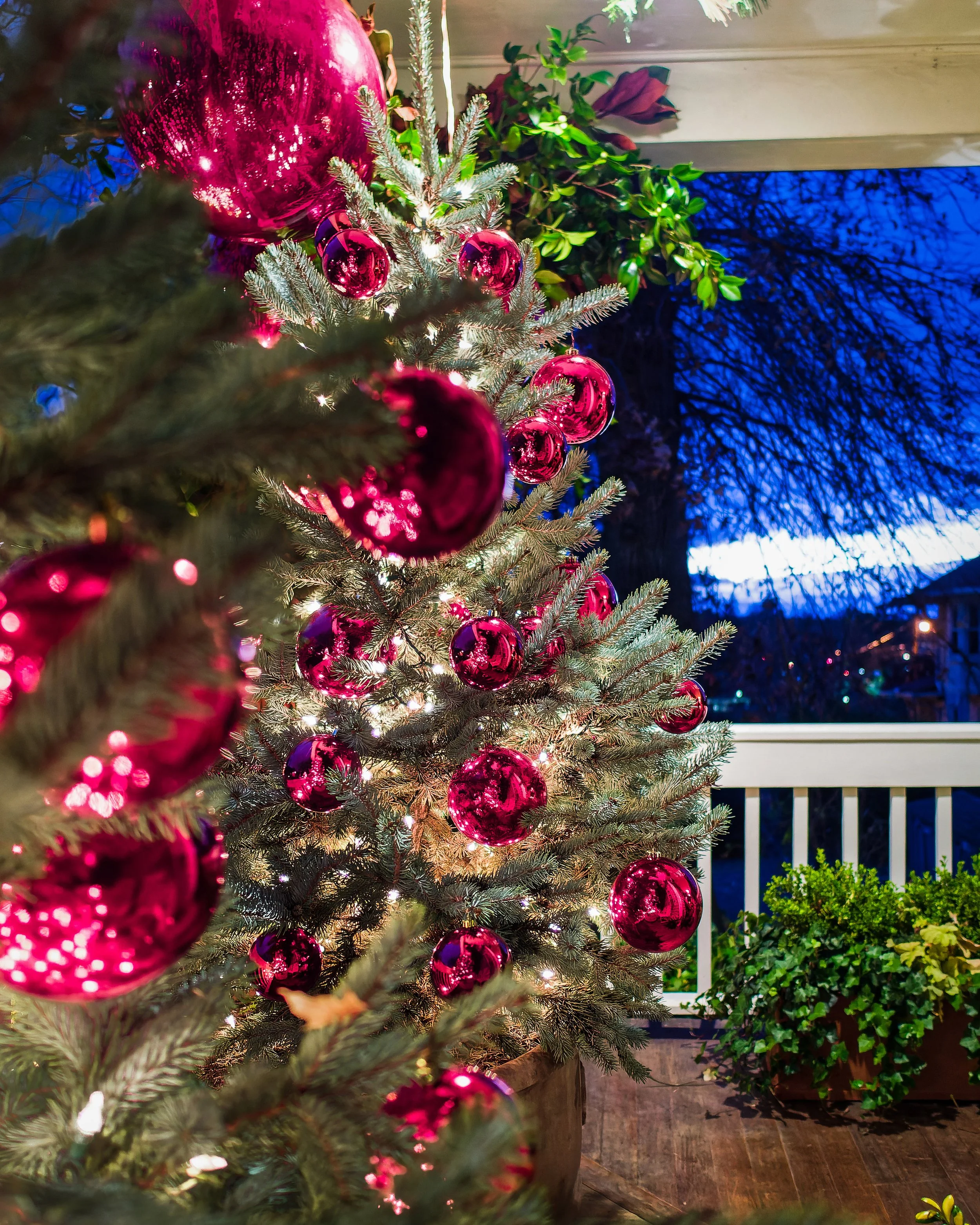 Decorated Christmas tree with pink ornaments on an outdoor porch at dusk, with a large tree and houses visible in the background.