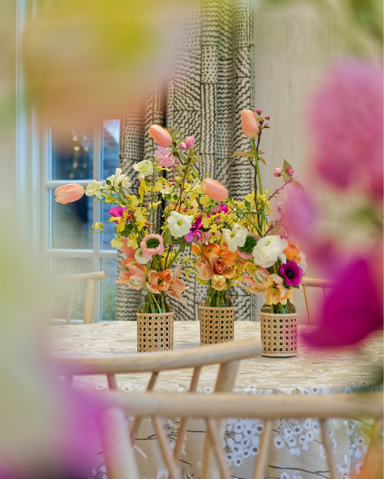 Three vases with colorful flower arrangements on a wooden table in a room with a window, curtains, and chairs, viewed through a blurred floral foreground.