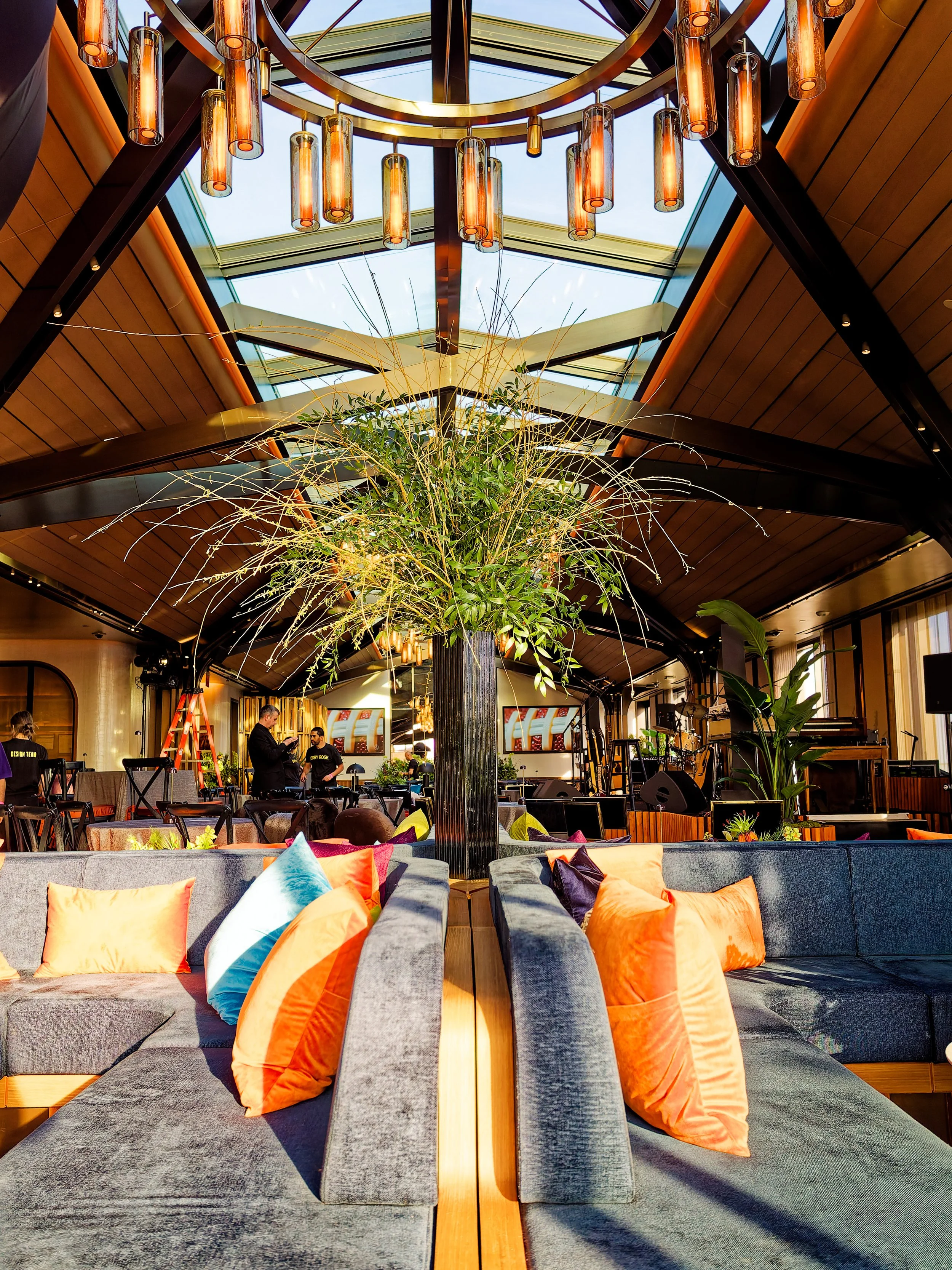 Interior of a modern venue featuring a large central plant and colorful pillows on grey couches, with a high glass ceiling, decorative lighting, and a stage area with instruments.