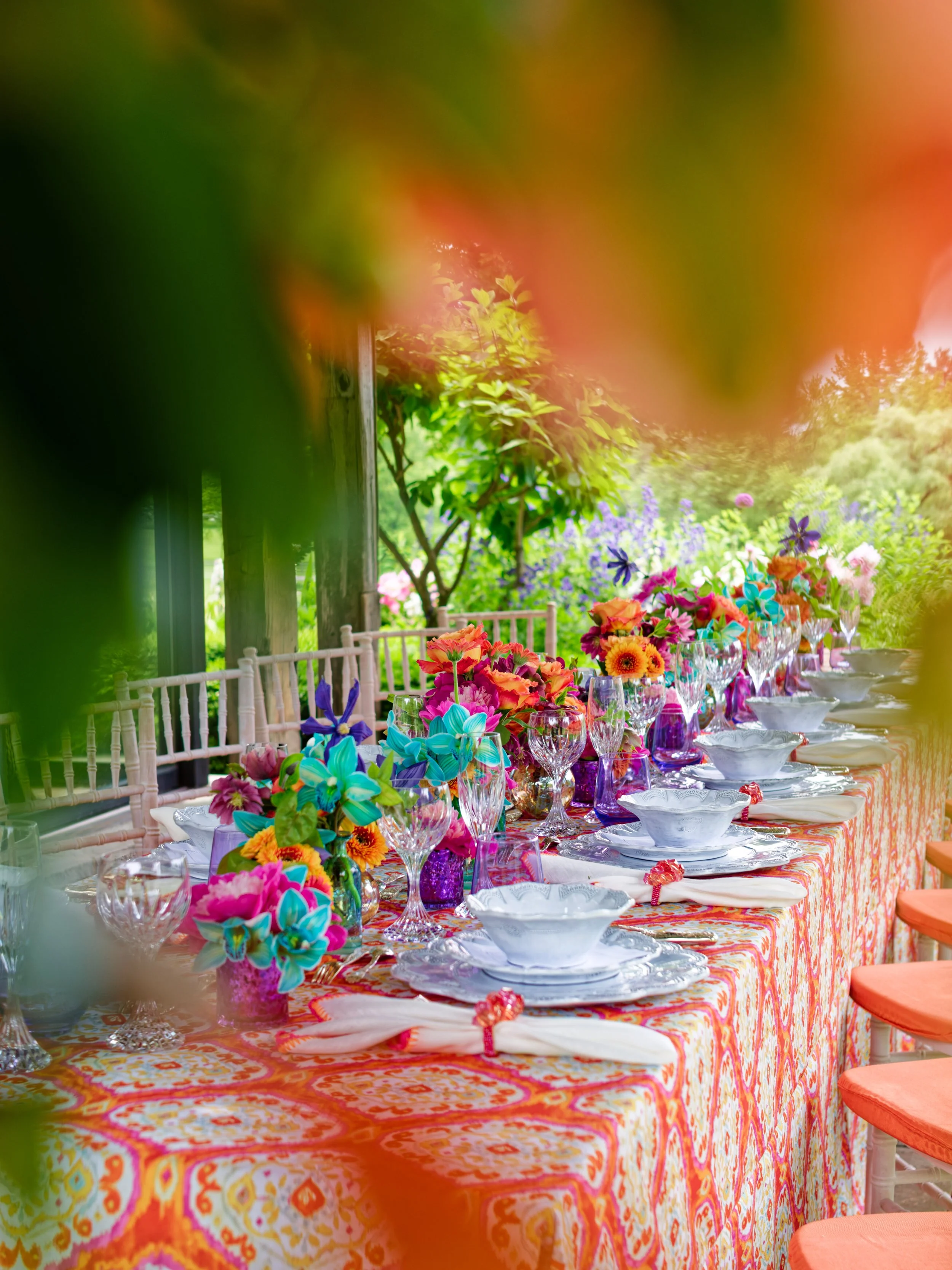 Colorful outdoor table setting with floral arrangements, glassware, and decorated plates on a brightly patterned tablecloth, surrounded by lush greenery and vibrant flowers.