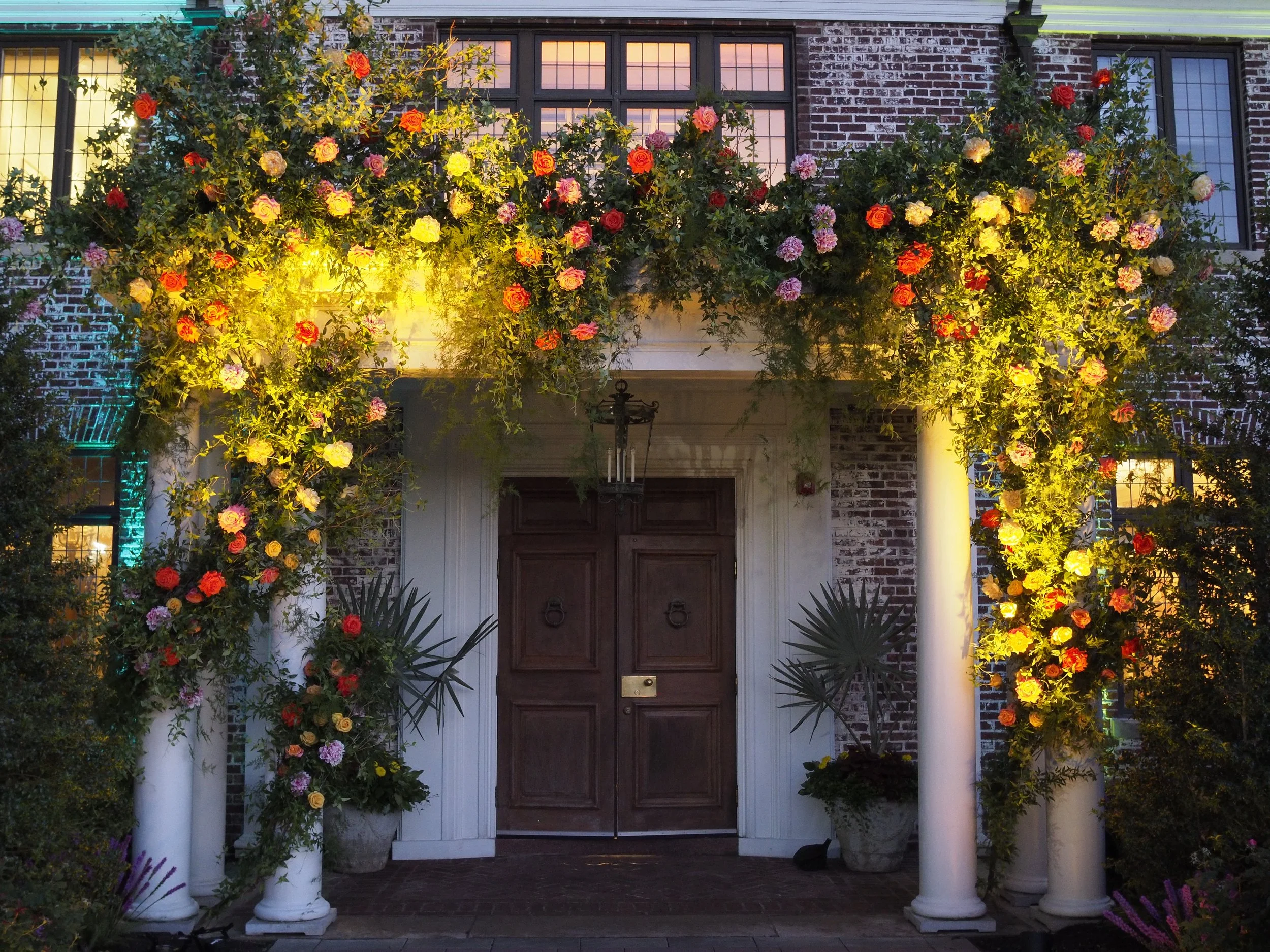 A house entrance decorated with a large arch of colorful flowers and greenery, illuminated with warm yellow lighting, including potted plants and brick walls.