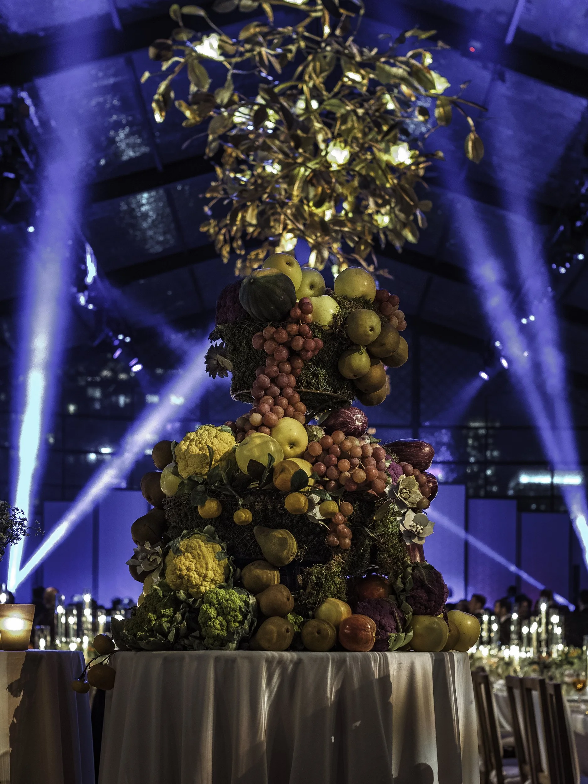 An elaborate floral centerpiece made of various fruits and flowers at a formal event with blue lighting and a tent-like ceiling in the background.