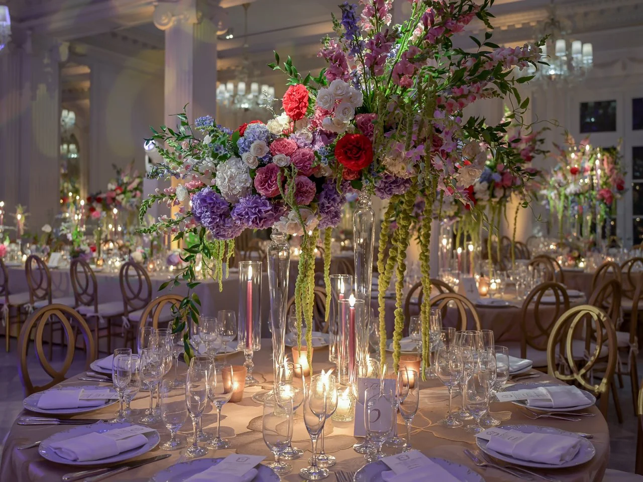 Elegant banquet table decorated with large floral centerpieces featuring pink, purple, red, and white flowers, surrounded by candles and glassware in a well-lit reception hall.