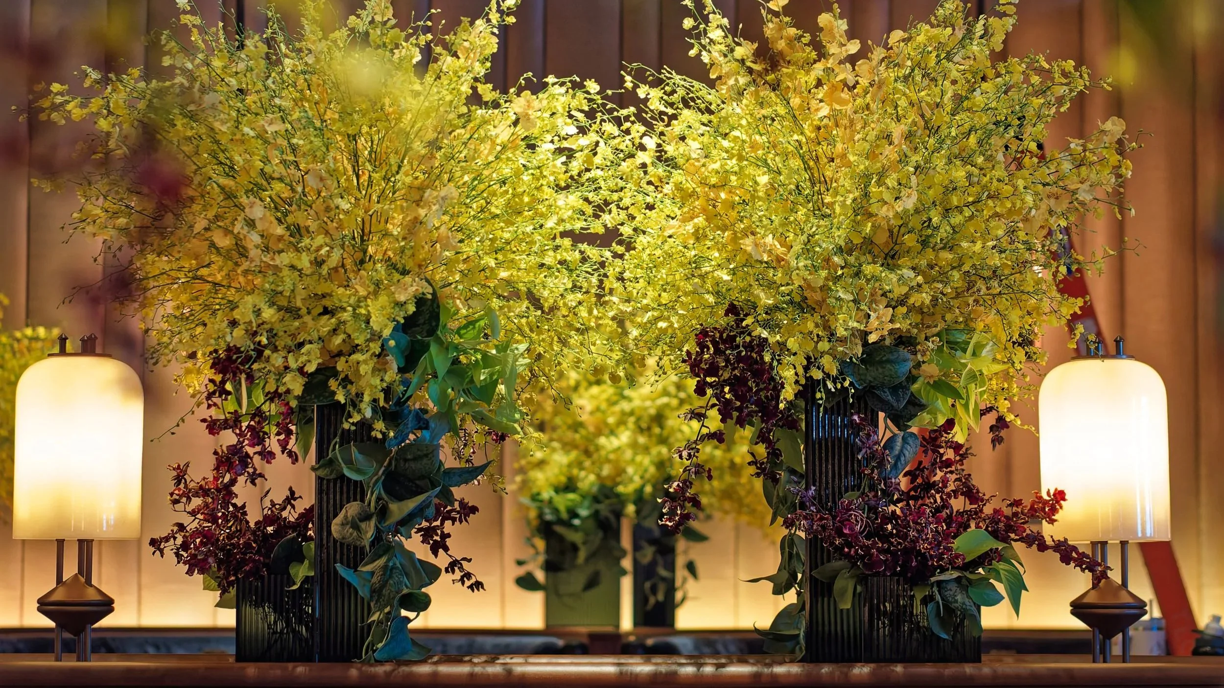 Floral arrangements with yellow and purple flowers in black vases, flanked by modern table lamps with white shades, against a warm decorated interior background.