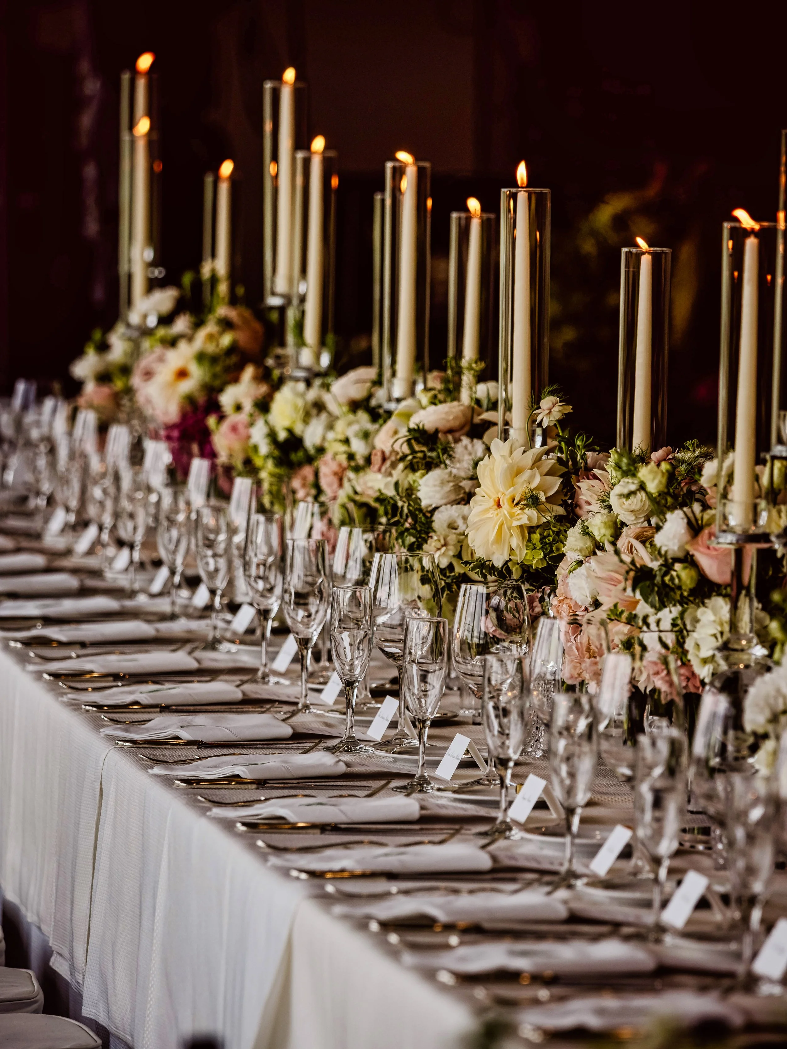 An elegant banquet table decorated with tall white candles, floral arrangements with white and pink flowers, many water glasses, and neatly folded white napkins.