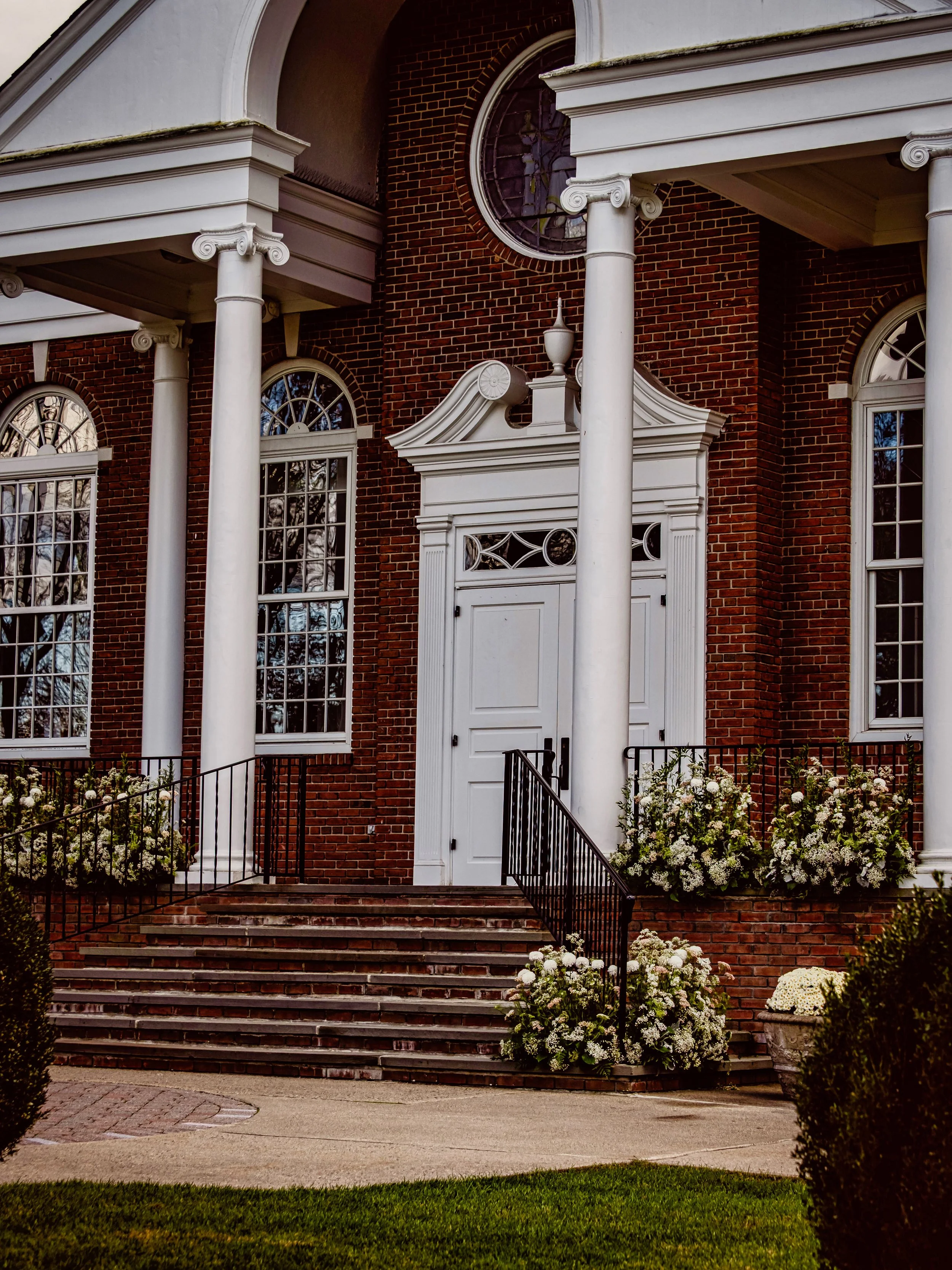 Front view of a brick house with white columns, stairs leading to a white door, large windows, and flower arrangements.