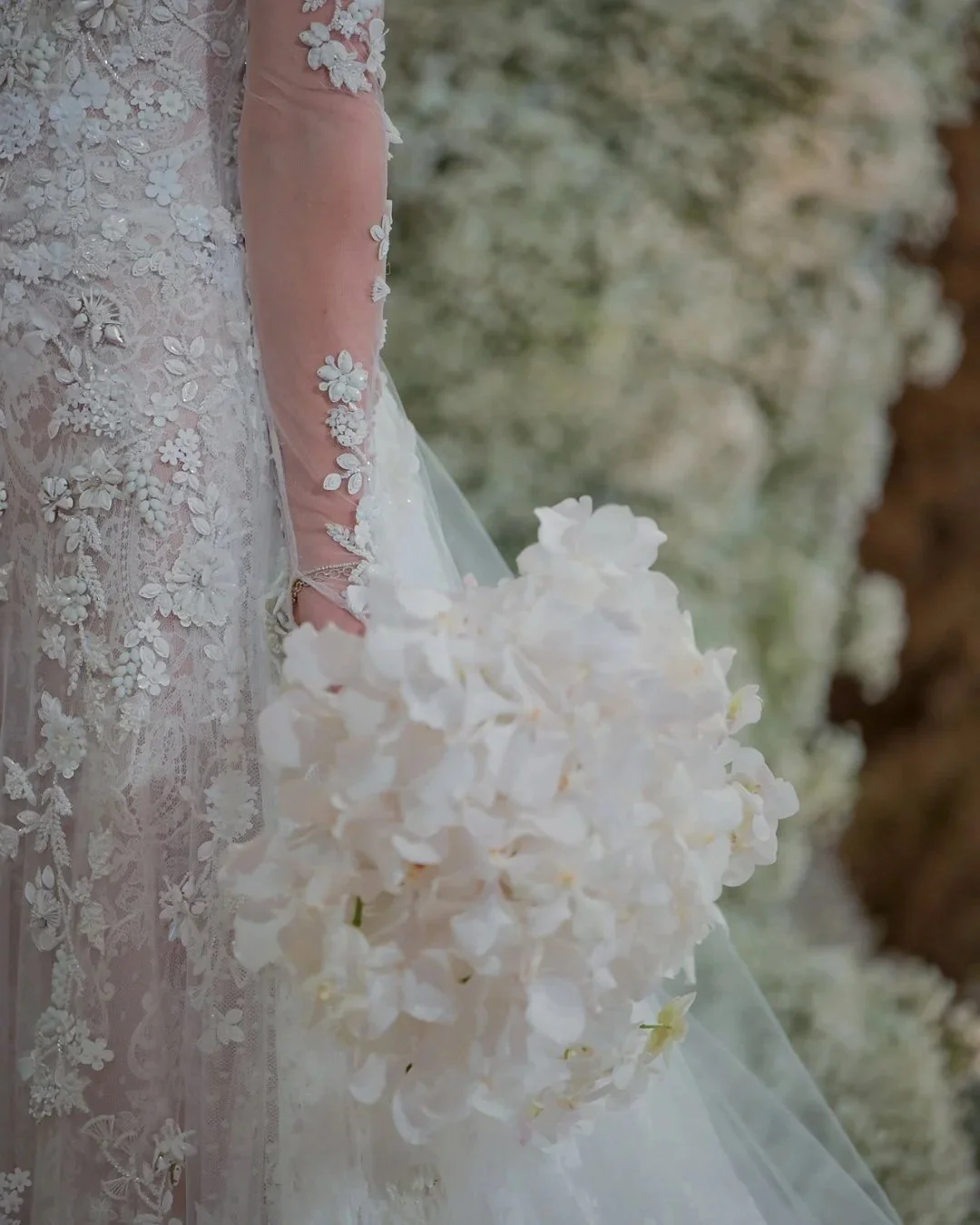 Part of a bride’s lace wedding gown with floral embroidery, holding a bouquet of white flowers against a blurred floral background.