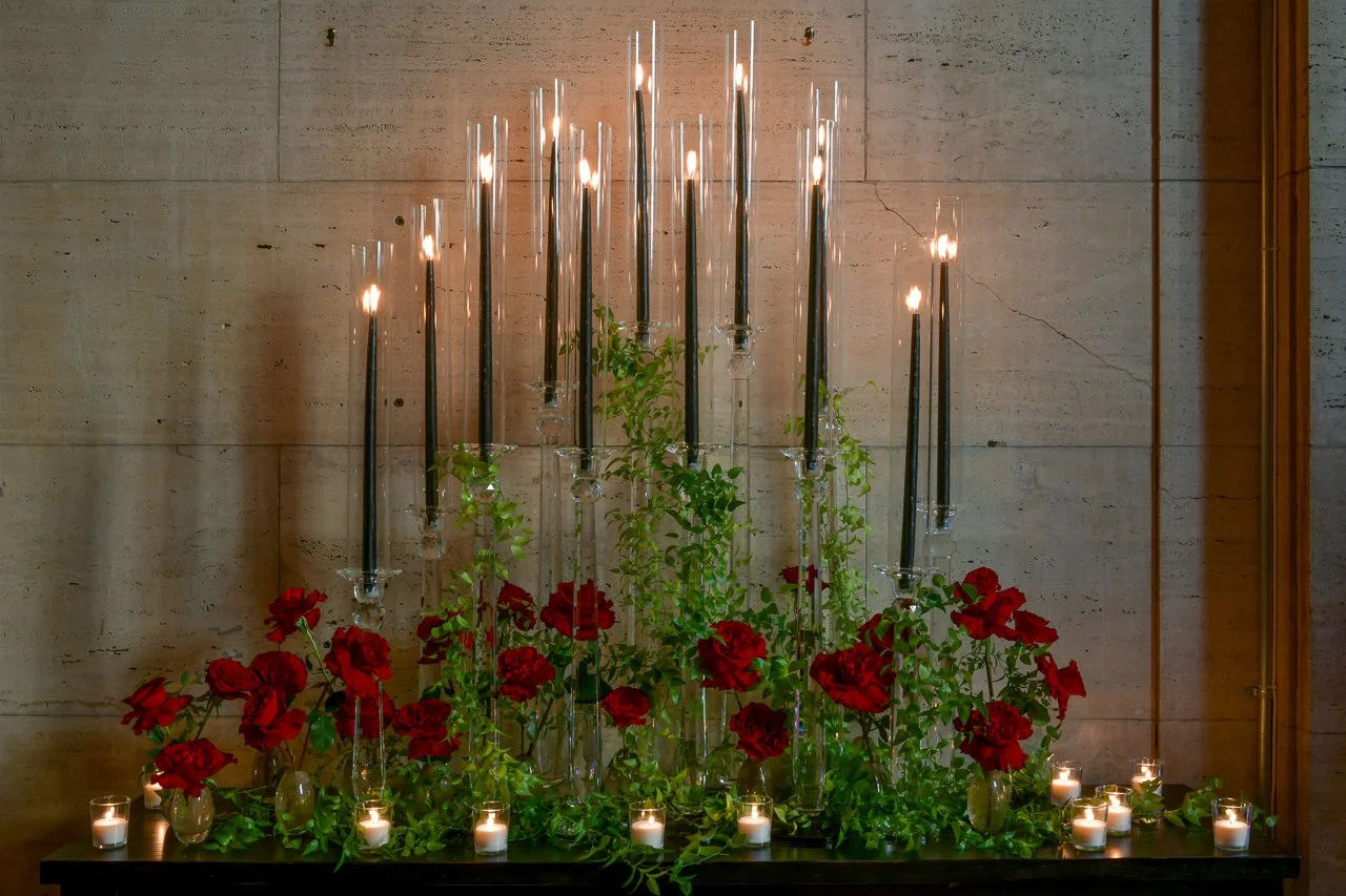 Decorative display with tall black candles in glass holders, surrounded by red flowers and small lit candles in glass containers on a table against a wooden wall.