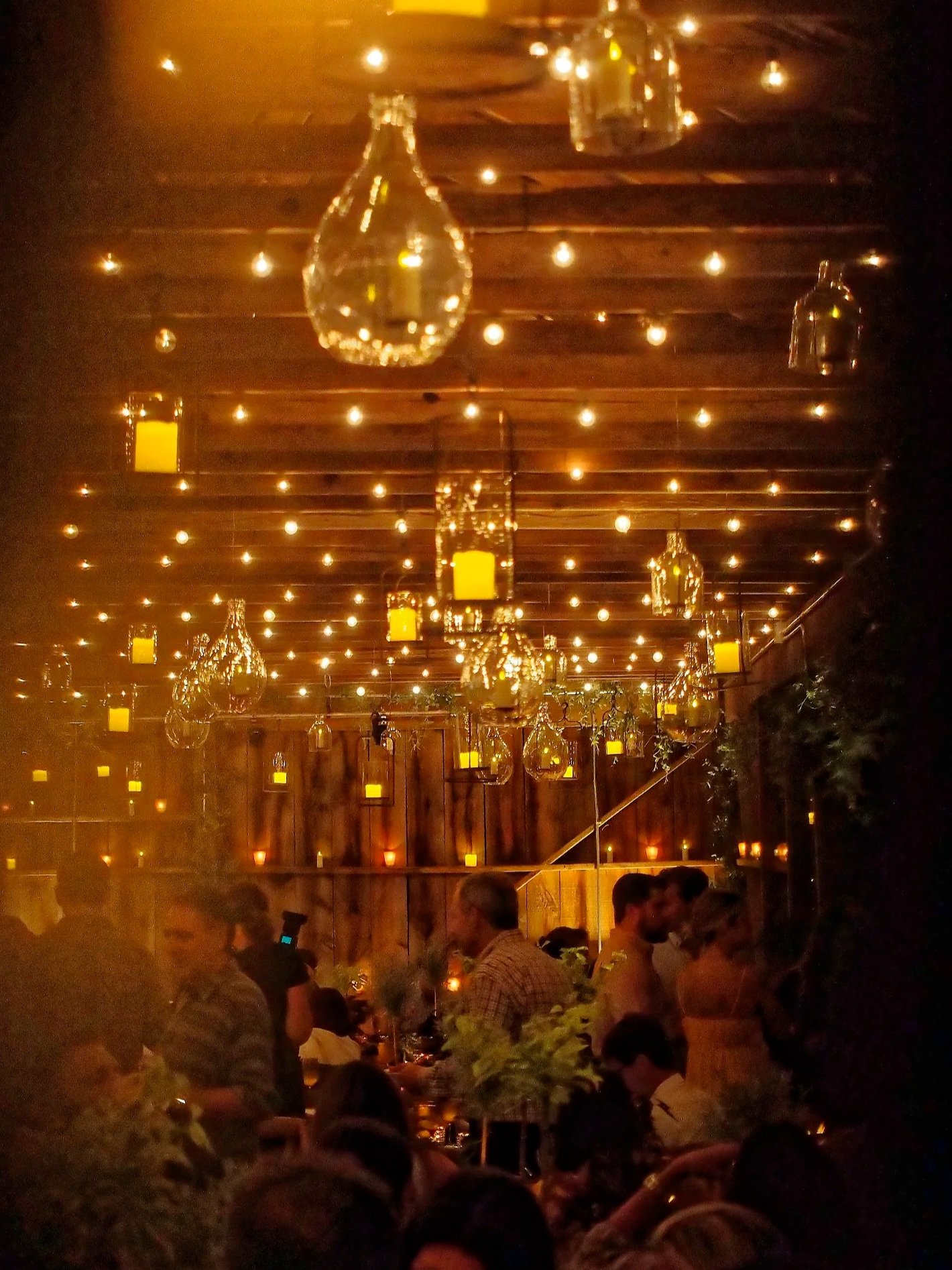 Dimly lit restaurant with hanging glass bottles and candles, many diners seated and socializing.