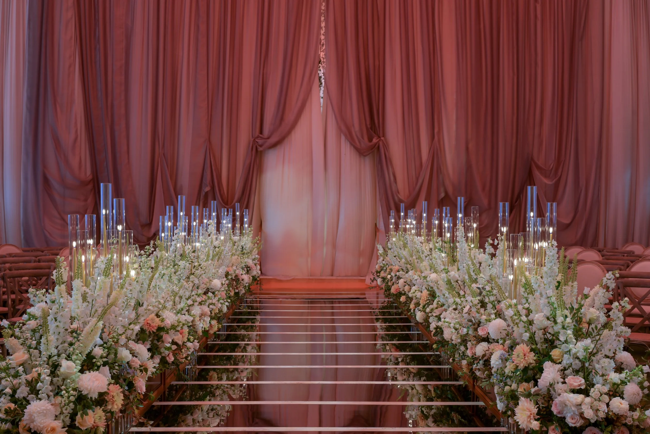 Elegant wedding aisle with pink drapes, floral arrangements, tall glass candles, and a mirrored walkway.