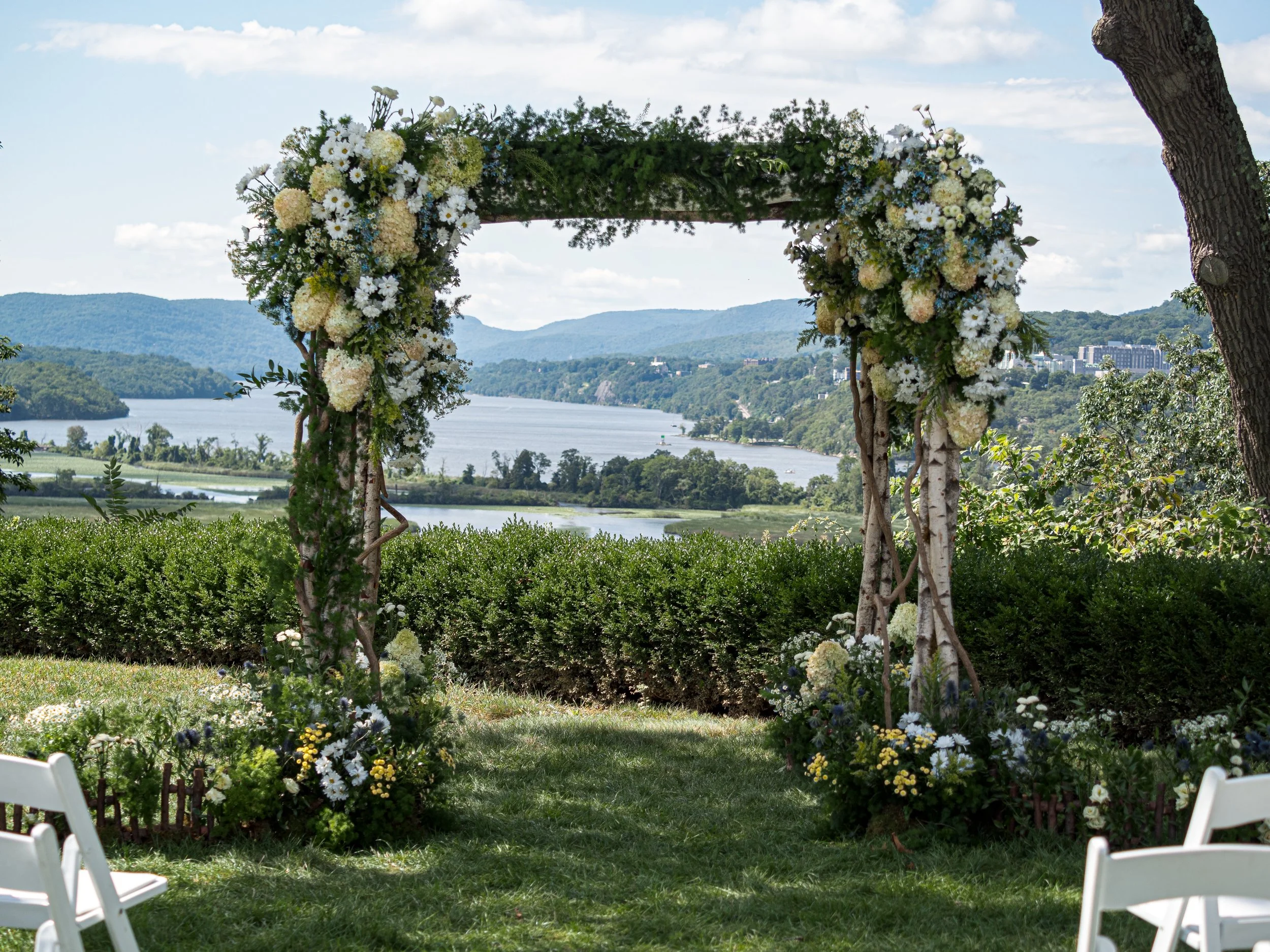 Outdoor wedding arch decorated with white and cream flowers, overlooking a river and distant hills, with white chairs in the foreground.