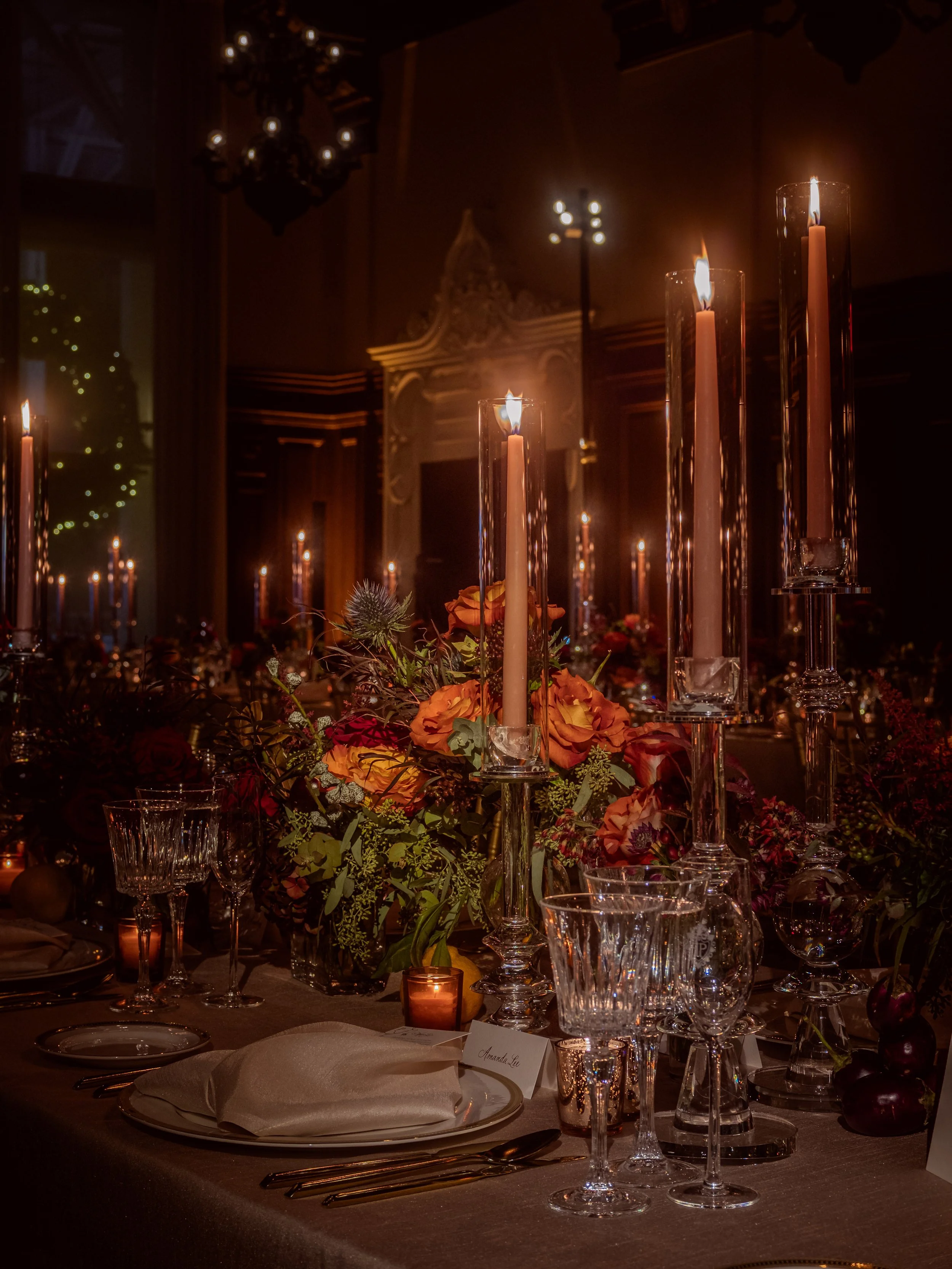 Elegant dinner table setting with candles, floral arrangements, glassware, and place cards in dim lighting.
