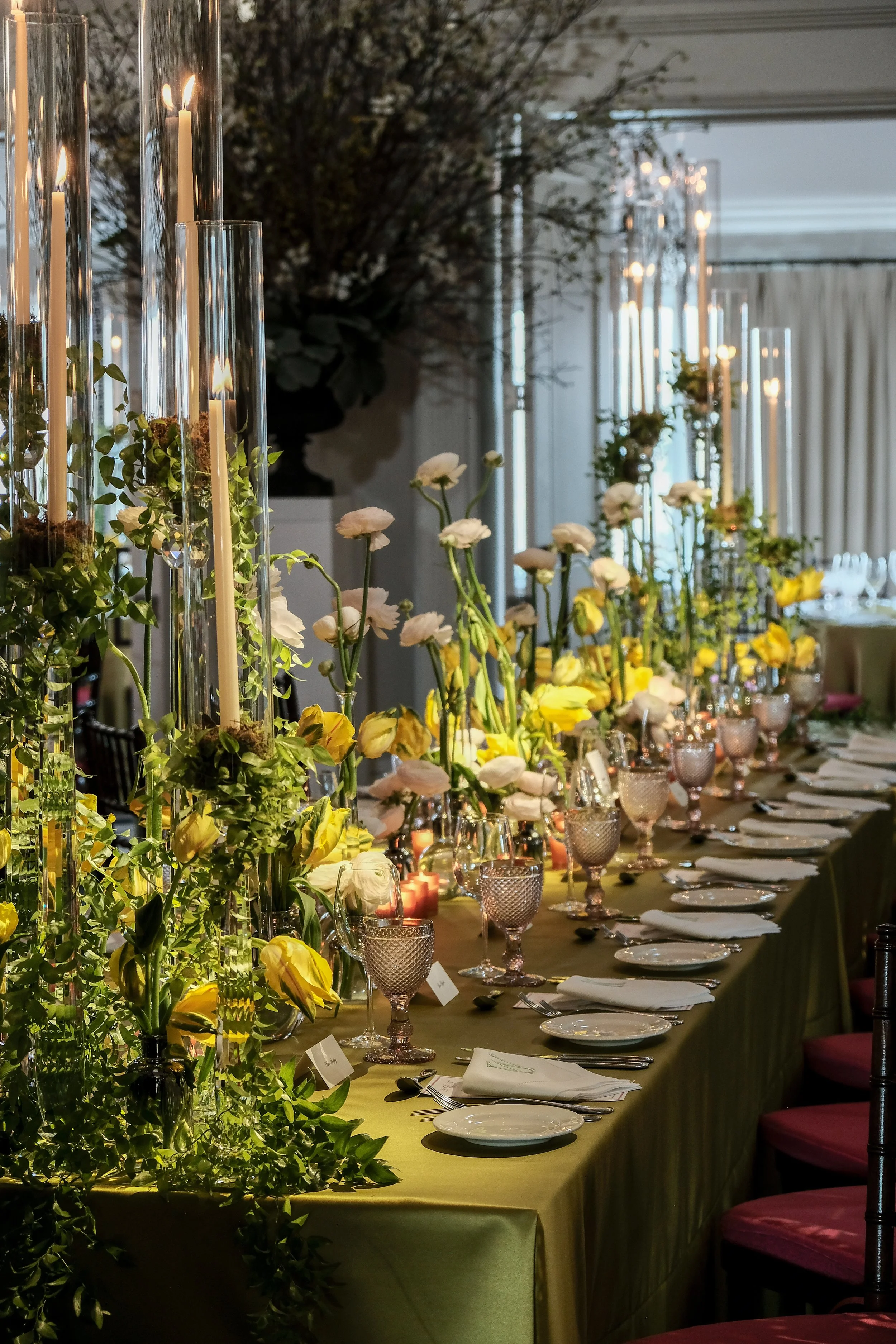 Elegant banquet table set with white and yellow flowers in tall glass vases, candles, and fine tableware, in an upscale event setting.