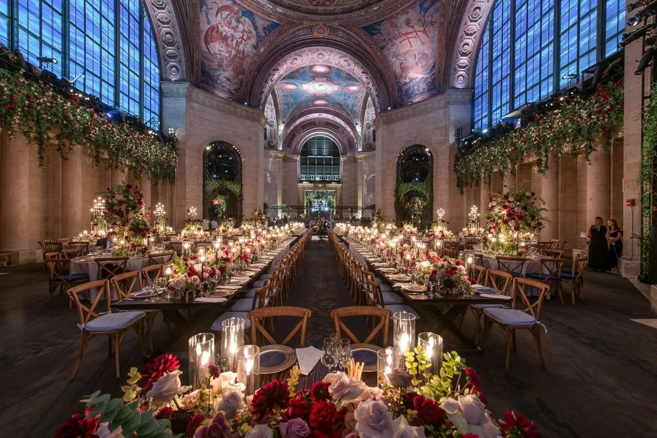 Elegant banquet hall decorated for a wedding or special event with long tables, floral arrangements, candles, and ornate ceiling art, illuminated by natural light from large windows.