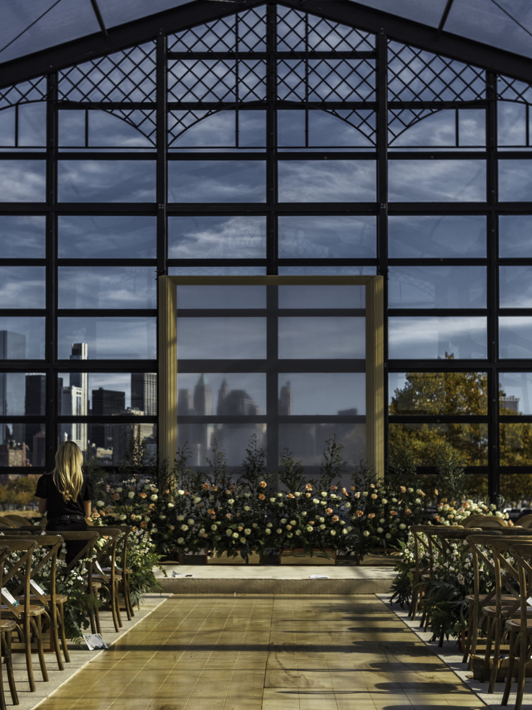 Indoor event space with a large glass wall overlooking a city skyline, decorated with flowers and chairs arranged for a ceremony.