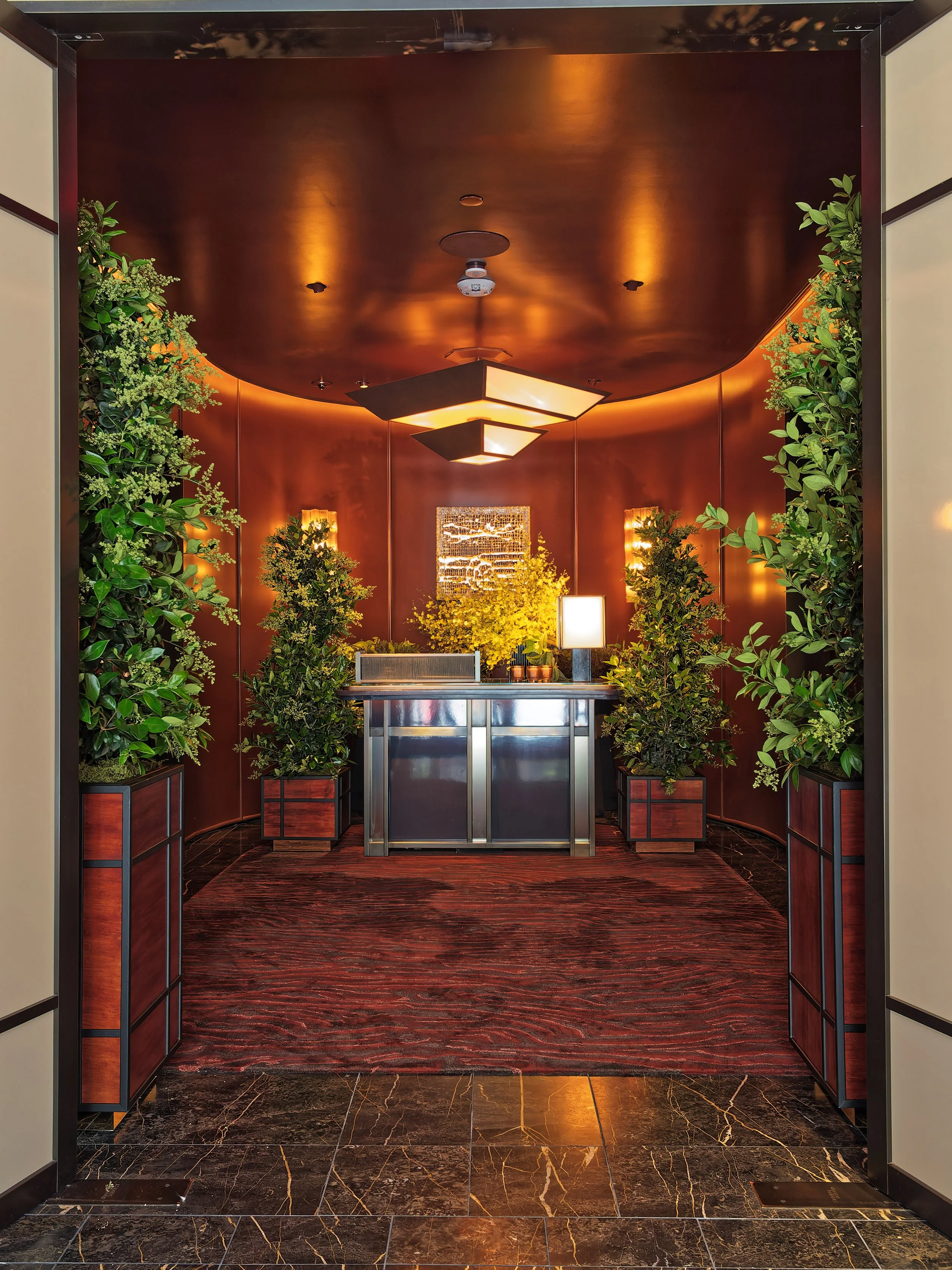 Elegant hotel lobby with a reception desk, surrounded by green plants in wooden planters, warm lighting, and a rich wooden ceiling and walls.