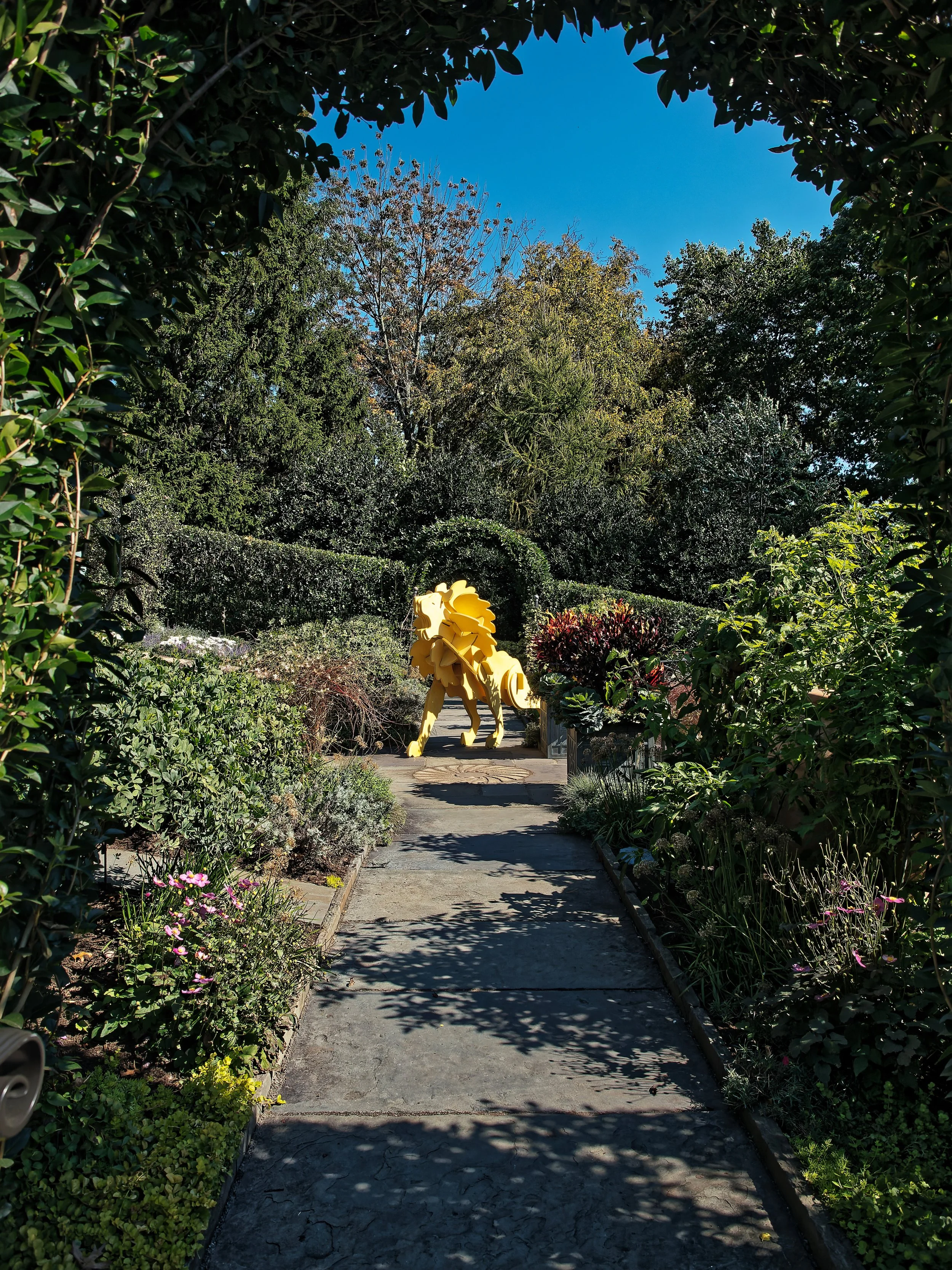 Pathway through a lush garden with various plants and flowers, leading to a large yellow lion sculpture at the end of the path, under a clear blue sky.