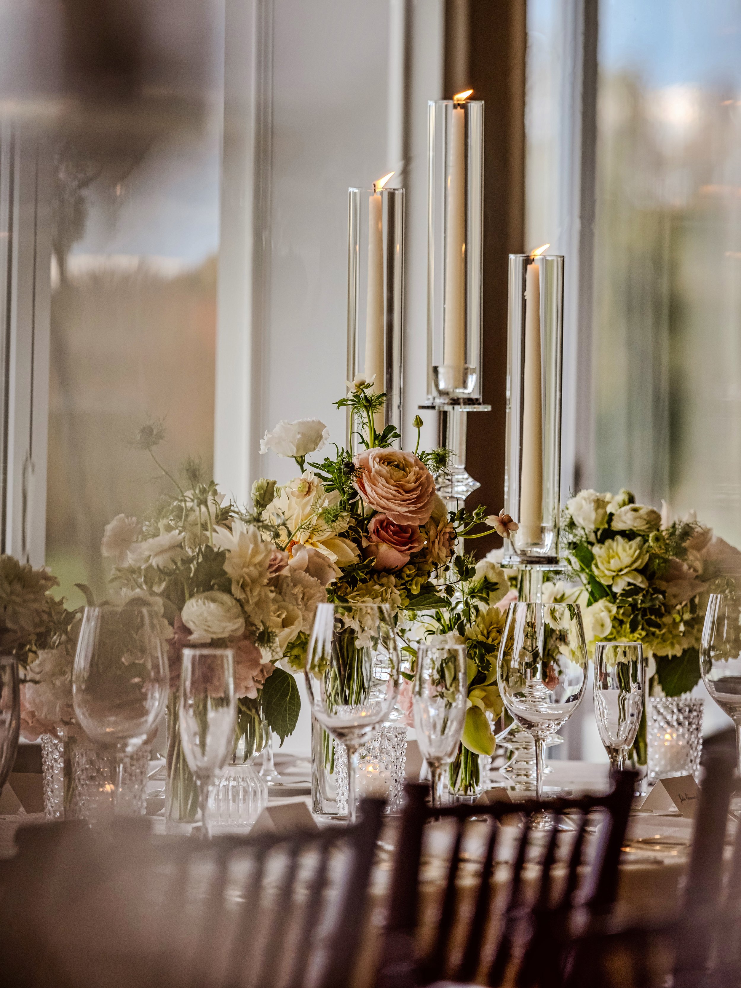 Elegant table centerpiece with pastel flowers, tall candles in glass holders, and crystal glassware, near a window with natural light.