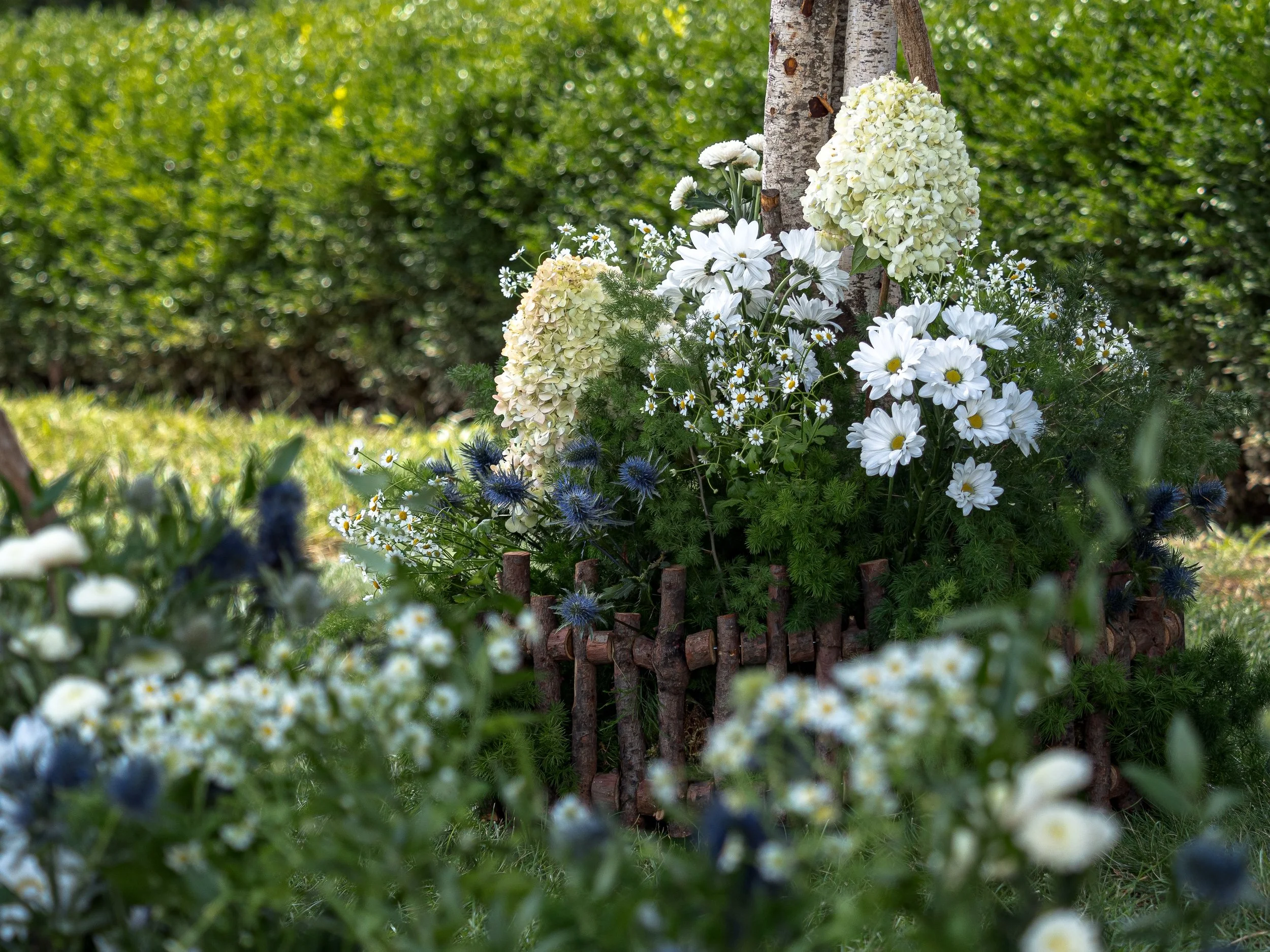 A garden bed filled with white hydrangeas, daisies, and other flowers, surrounded by an artificial wooden fence with a tree trunk behind it, and a background of greenery.