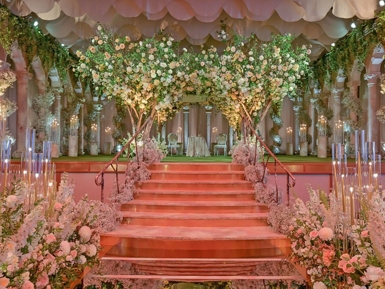 Elegant wedding altar decorated with abundant pink and white flowers, including large trees and cascading floral arrangements, set on a raised platform with a pink staircase.