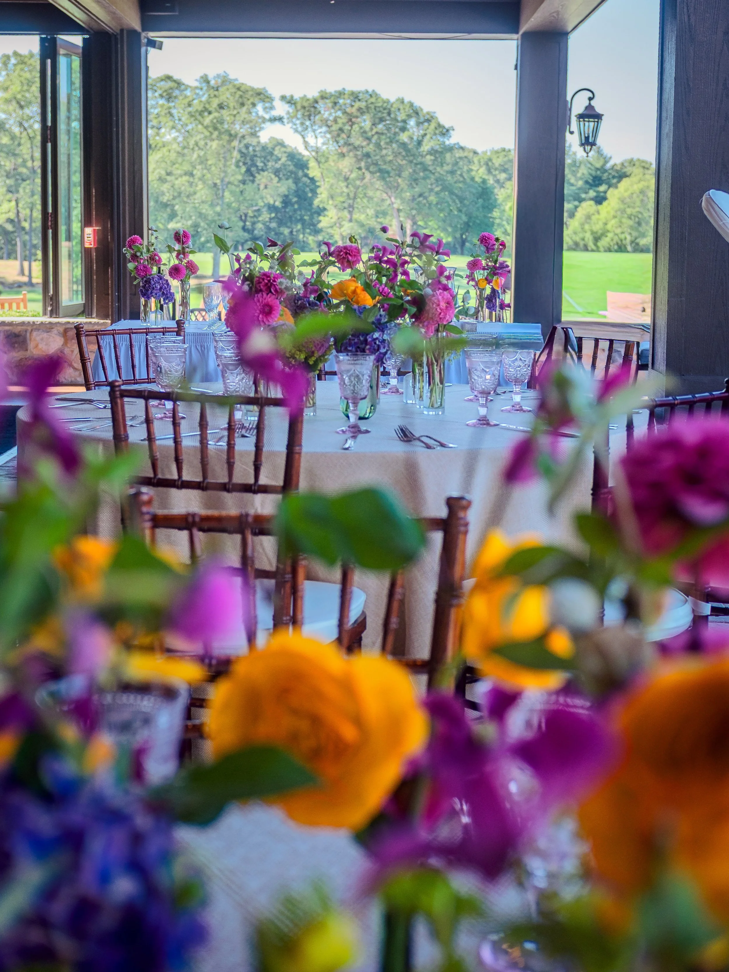 Banquet table decorated with colorful flowers and glassware inside a room with large open windows view of a green outdoor landscape.