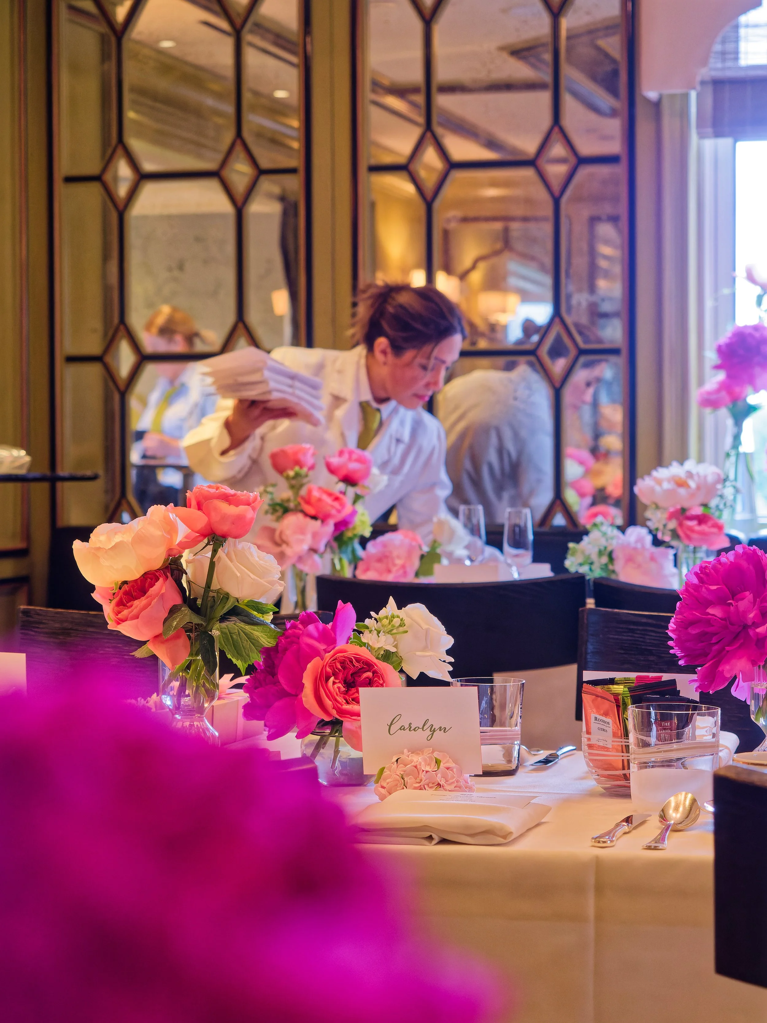 A woman setting a dinner table in a decorated banquet hall with pink and white floral centerpieces and a reflective geometric-patterned mirror on the wall.