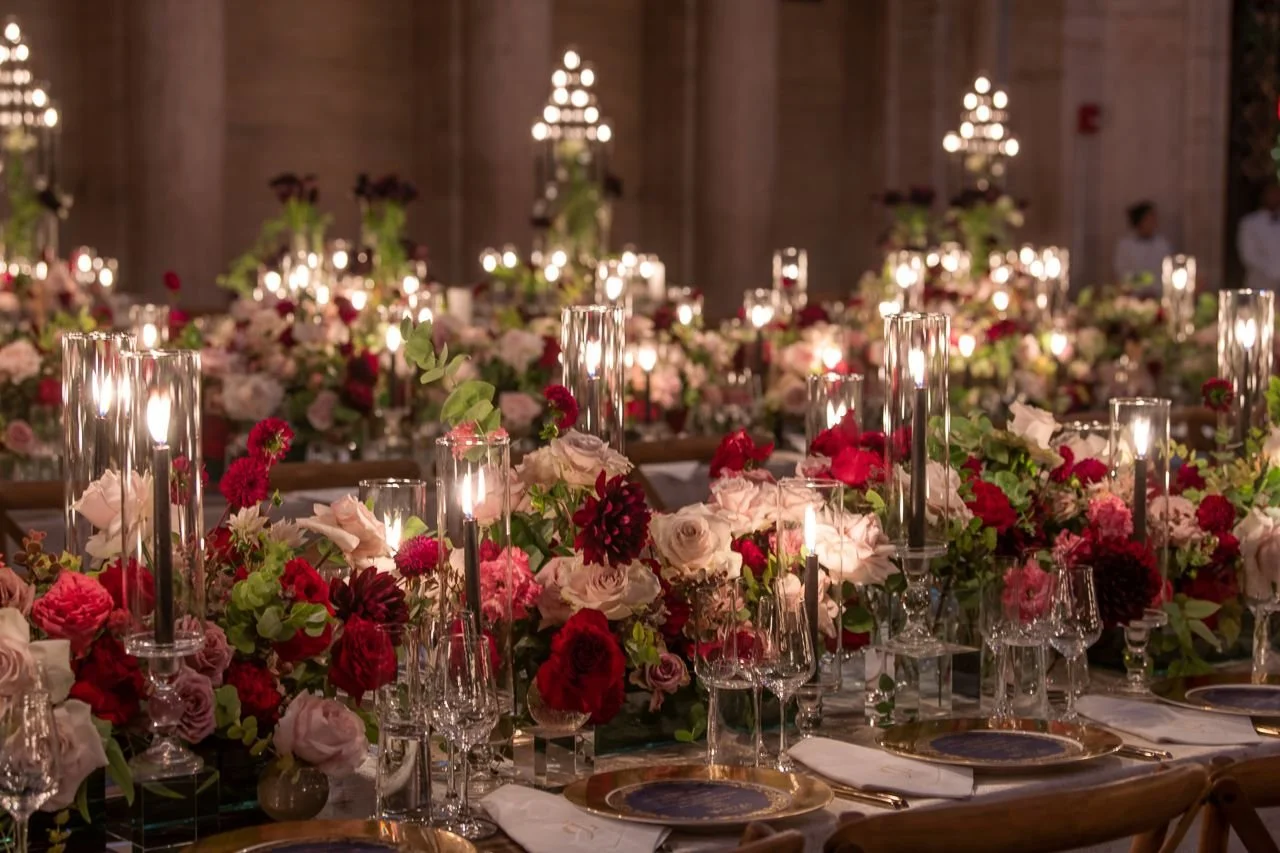 Elegant event table decorated with numerous pink, red, and white flowers and tall glass candle holders with lit candles, set in a grand hall.