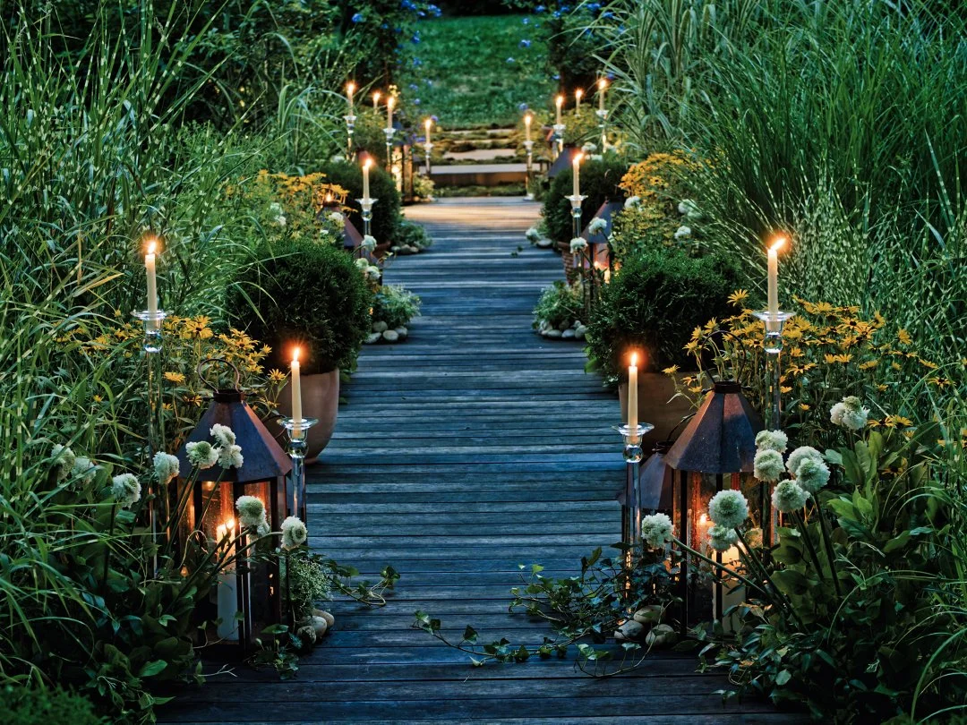 A wooden pathway decorated with lit candles and lanterns, surrounded by tall green grass, bushes, and yellow flowers.