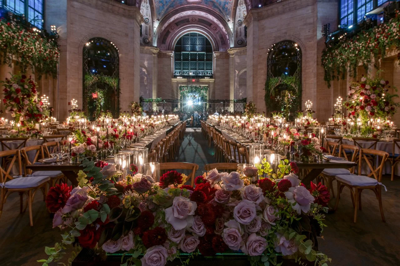 Elegant indoor wedding reception with long banquet tables decorated with candles, pink, red, and white flowers, and greenery. The venue has high arched ceilings, large windows, and ornate architectural details, creating a romantic and luxurious atmos