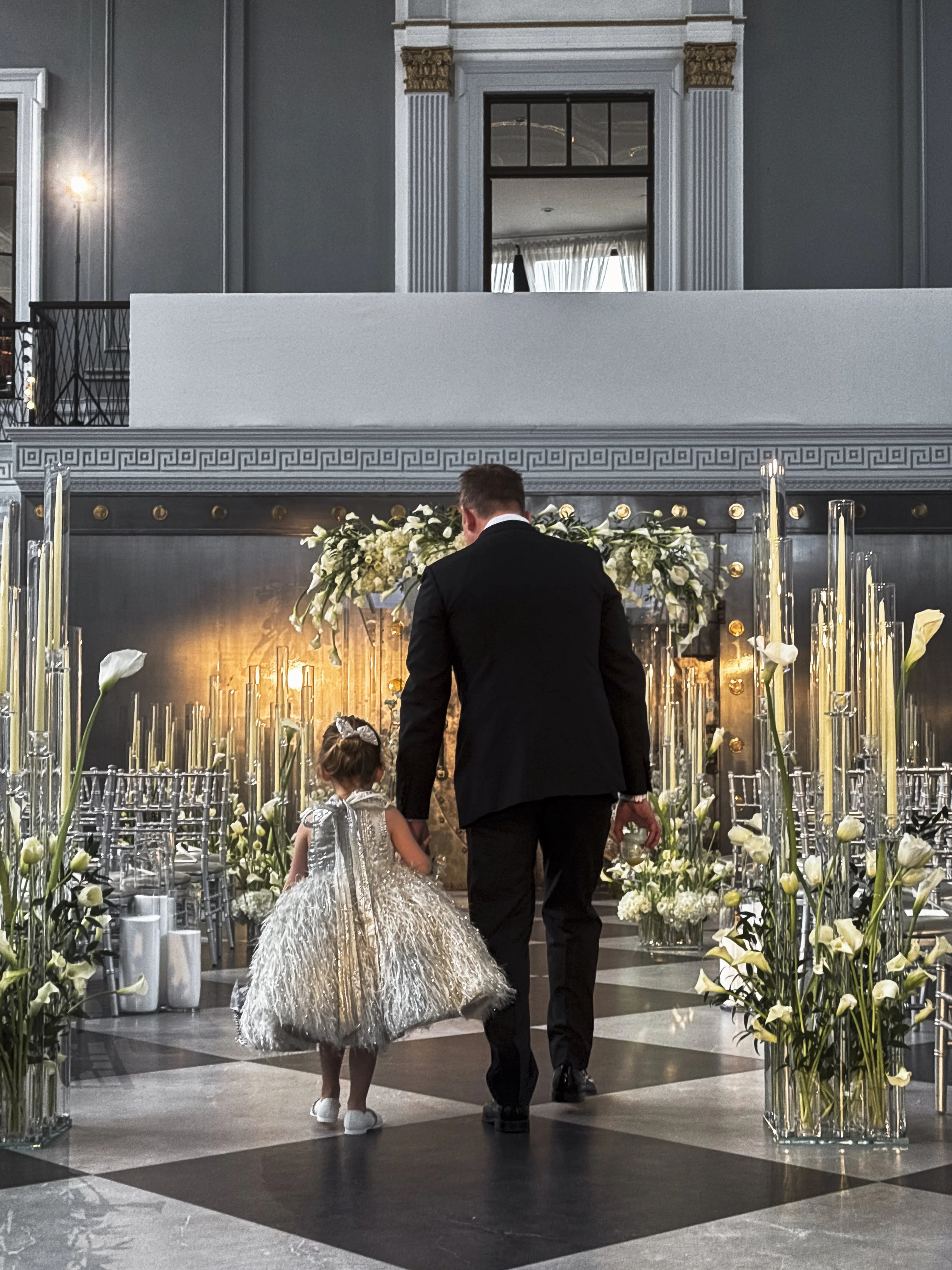 A man and a young girl walking hand in hand into a decorated event space with flowers and candle centerpieces, likely at a formal event.